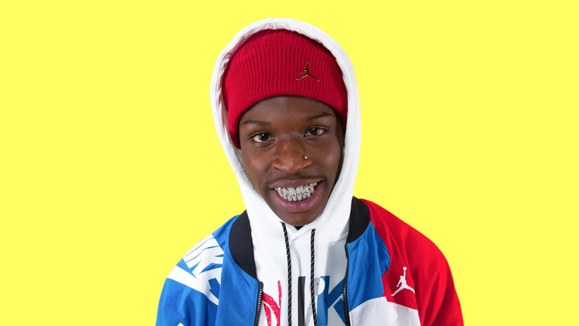 Quando Rondo 桌布 壁紙 — 一位年輕男子帶著燦爛笑容和鑽石牙套，戴著紅色毛帽，穿著色彩繽紛的Jordan連帽上衣，背景是鮮黃色。