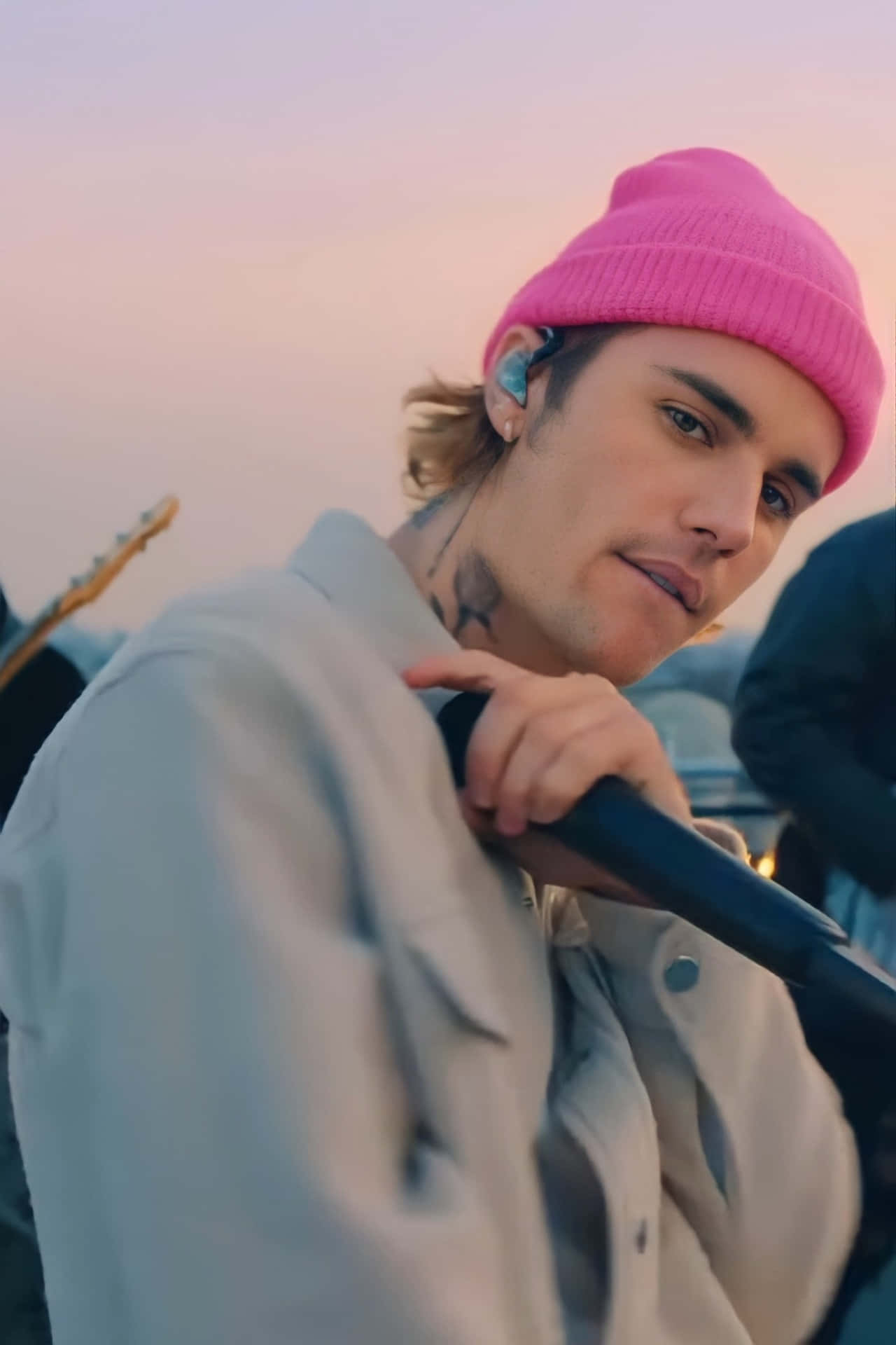 Justin Bieber 2021 桌布 壁紙 — 一位年輕男子戴著亮粉紅色毛帽和輕便外套，在柔和粉紅與橙色夕陽天空前拿著麥克風。
