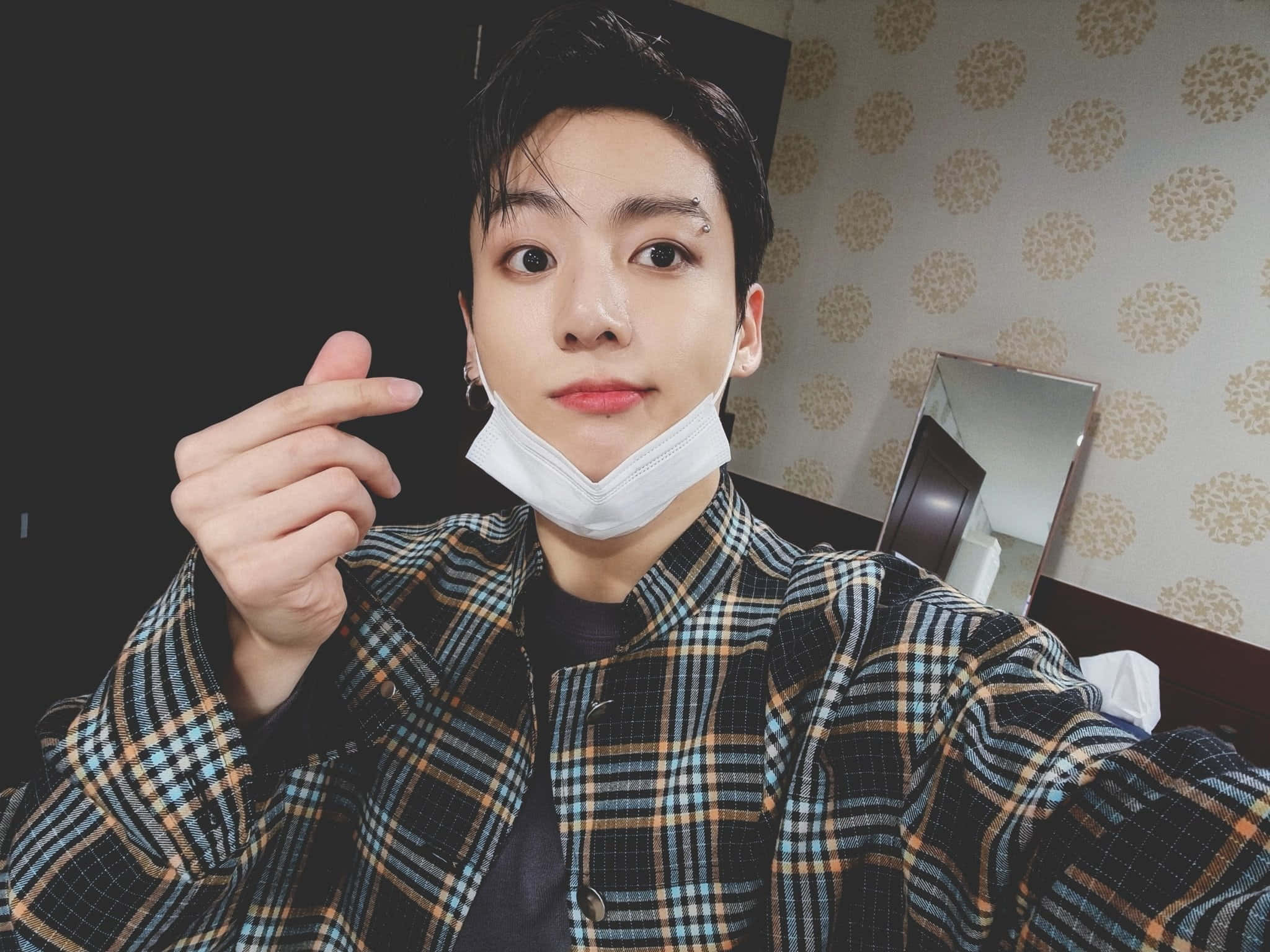 Jungkook 桌布 壁紙 — 一位年輕男子拉下口罩，微笑並指向相機，房間內有圖案壁紙。