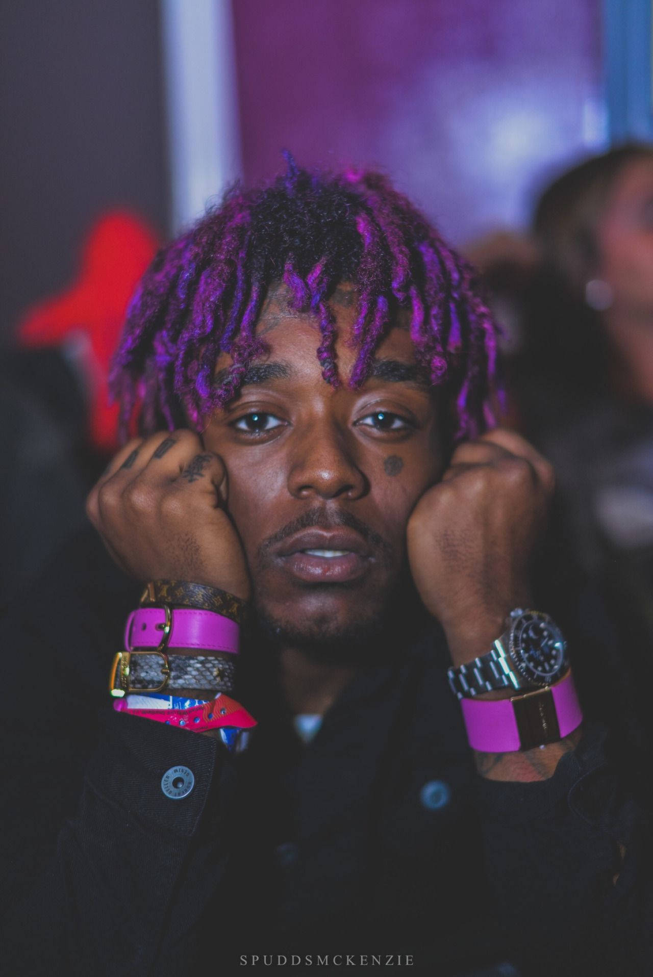 Uzi 桌布 壁紙 — 一位年輕男子擁有充滿活力的紫色辮子，將頭靠在拳頭上，在昏暗的俱樂部燈光下佩戴大膽的粉紅色和銀色飾品。