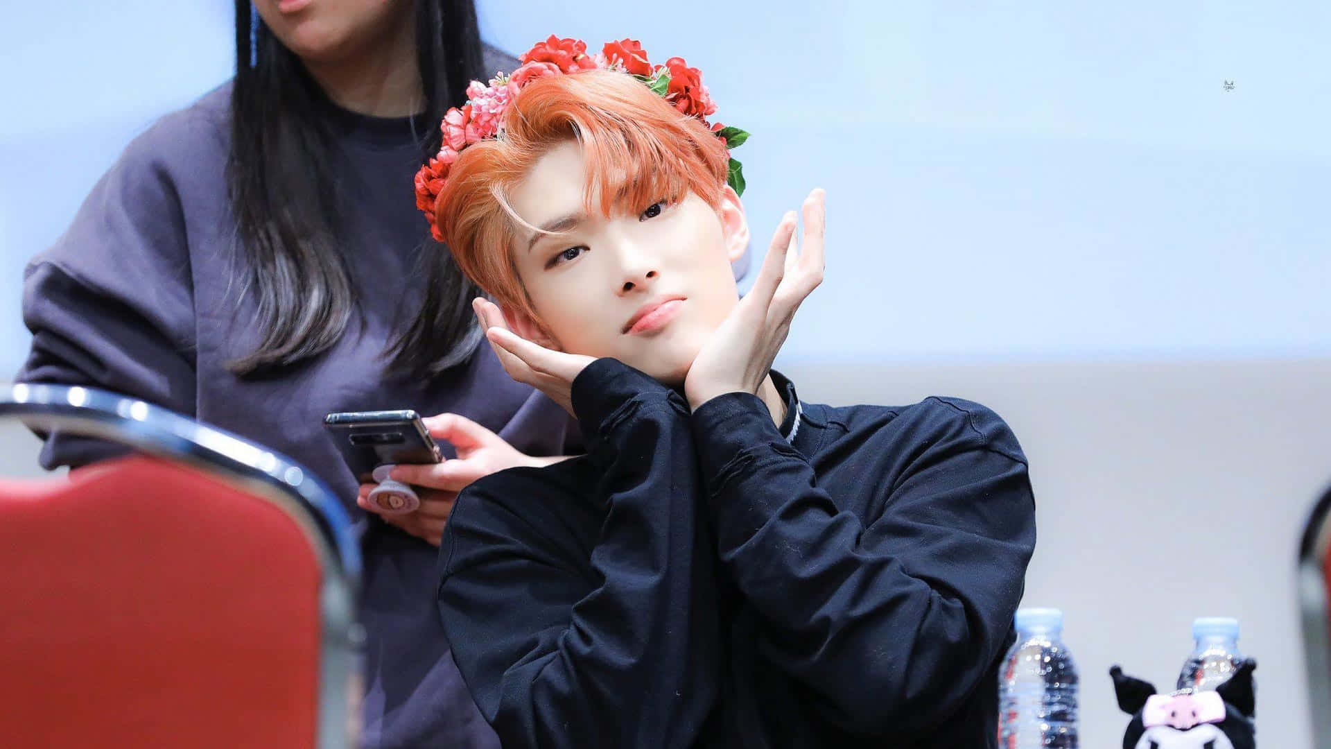 Ateez Desktop 桌布 壁紙 — 一位年輕男子擁有橙色頭髮，飾以紅色花朵，在明亮的室內環境中，雙手框著臉部，擺出俏皮的姿勢。