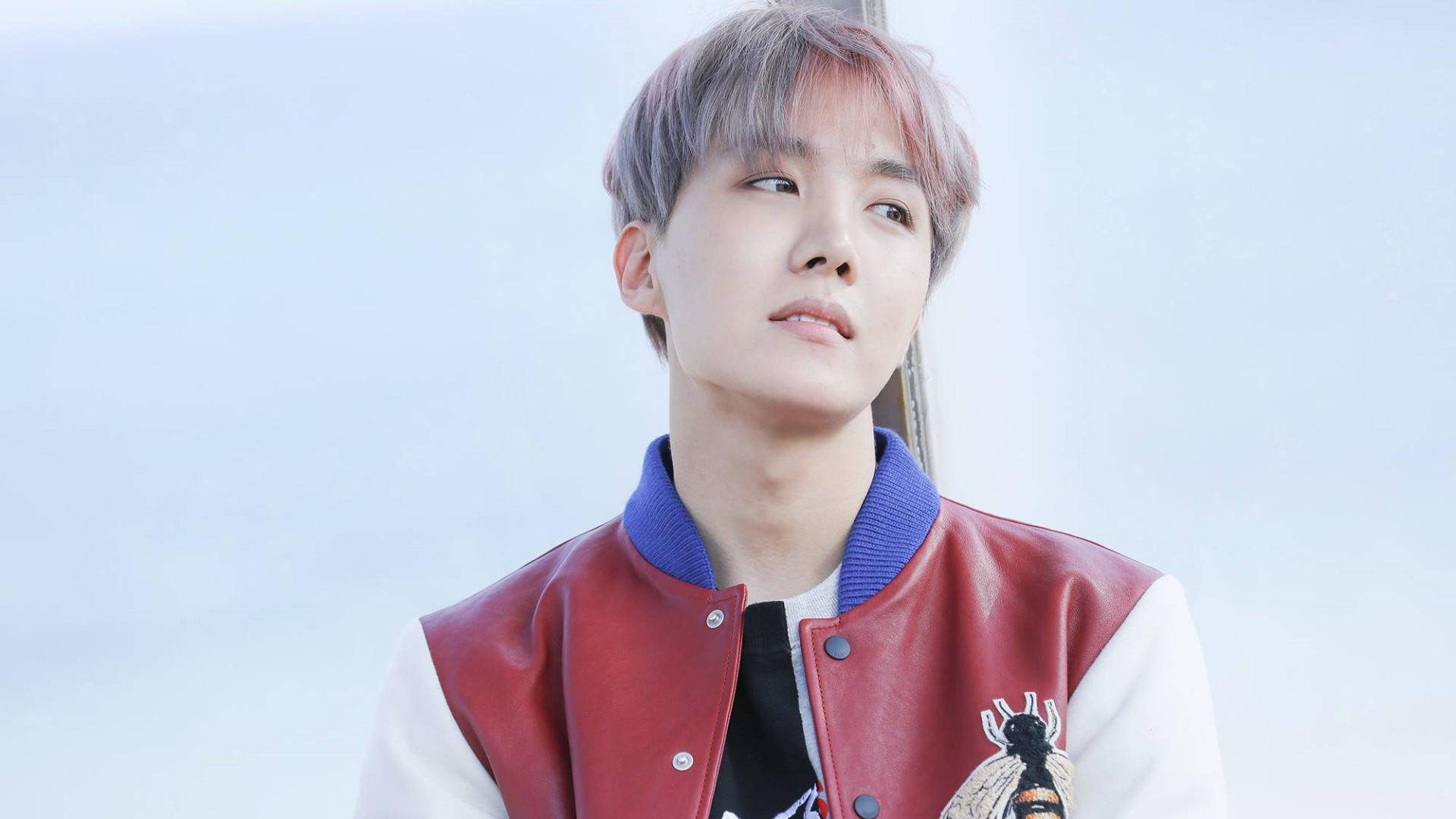 jhope 桌布 壁紙 — 一位年輕男子擁有粉彩粉紅和灰色頭髮，穿著紅白相間的校隊夾克，背景是柔和的天空。