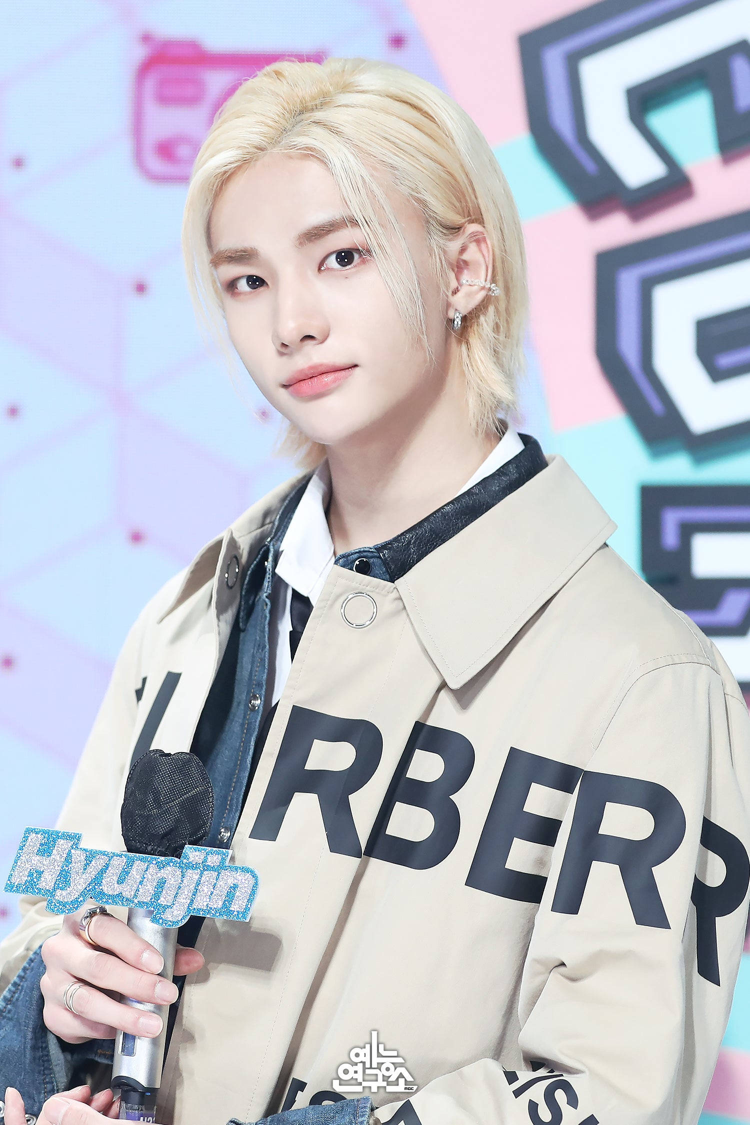 Stray Kids 桌布 壁紙 — 一位年輕男子擁有鉑金色頭髮，身穿米色Burberry外套，手持麥克風，背景為彩色幾何圖案。