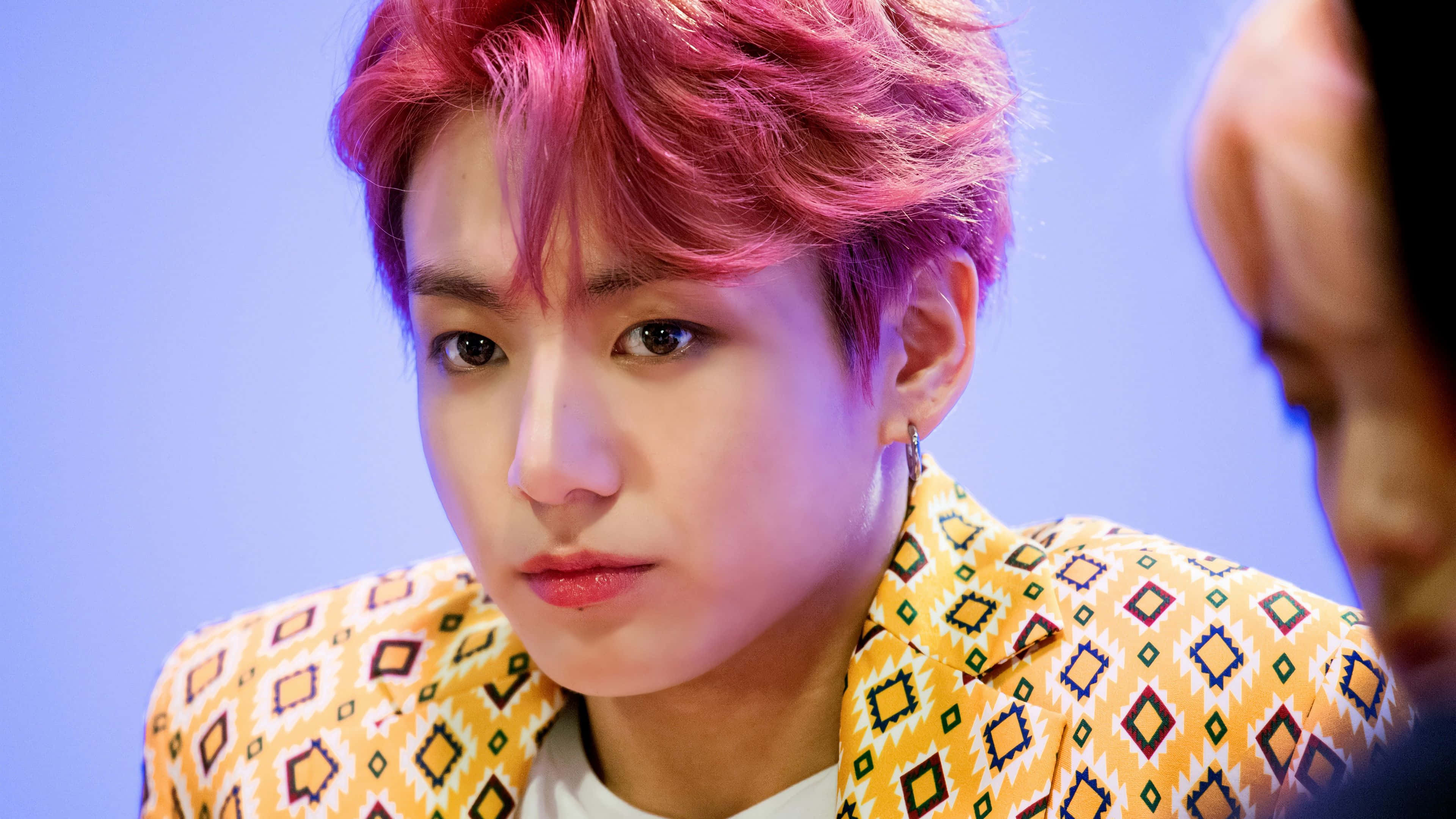Jungkook 桌布 壁紙 — 一位年輕男子擁有鮮豔的粉紅色頭髮，穿著大膽的黃色圖案西裝外套，背景是柔和的藍色。