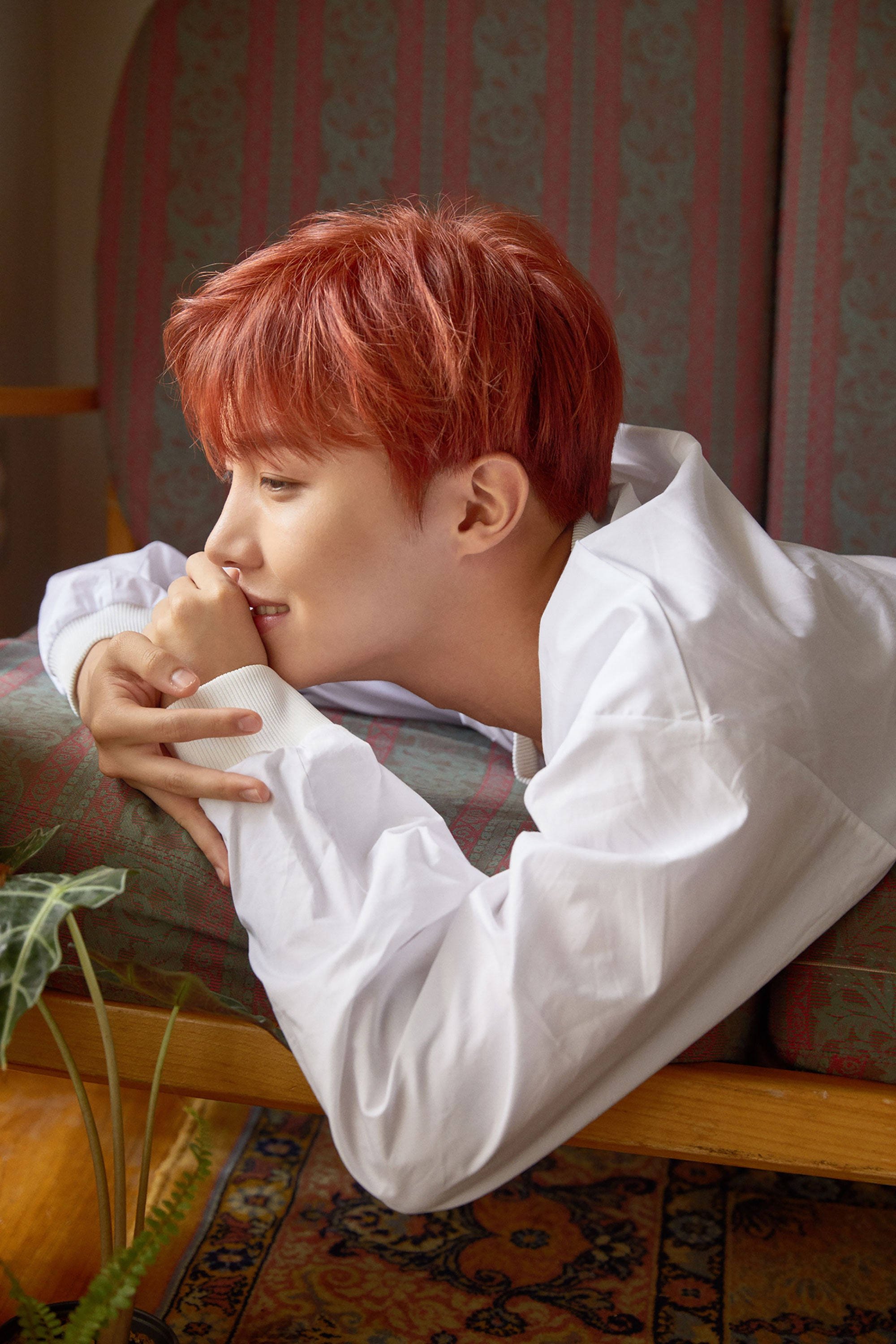 j-hope 桌布 壁紙 — 一位年輕男子擁有鮮豔的紅髮，在溫暖照明的房間裡，將下巴擱在交疊的雙手上，懶洋洋地躺在圖案沙發上。