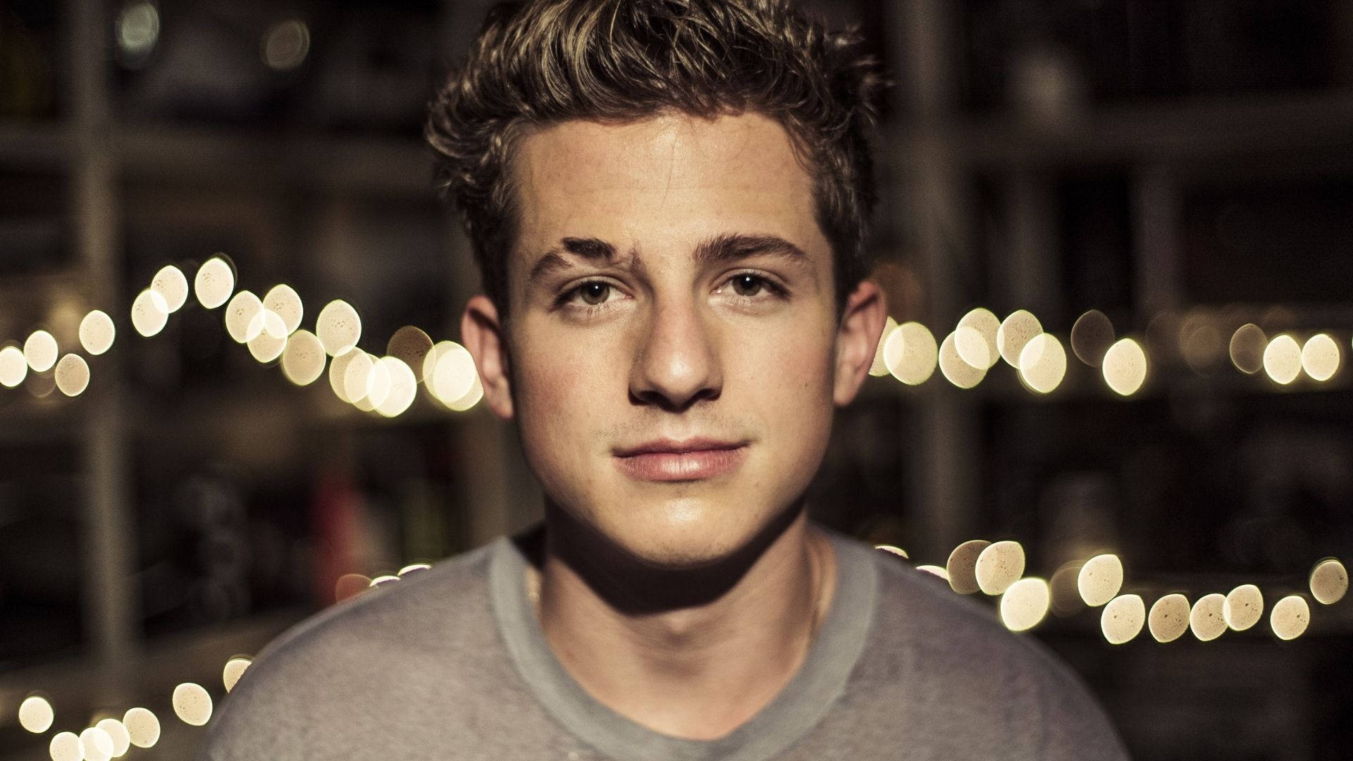 Charlie Puth 桌布 壁紙 — 一位年輕男子梳理著時尚髮型，在夜晚溫暖串燈的模糊背景下平靜地凝視鏡頭。