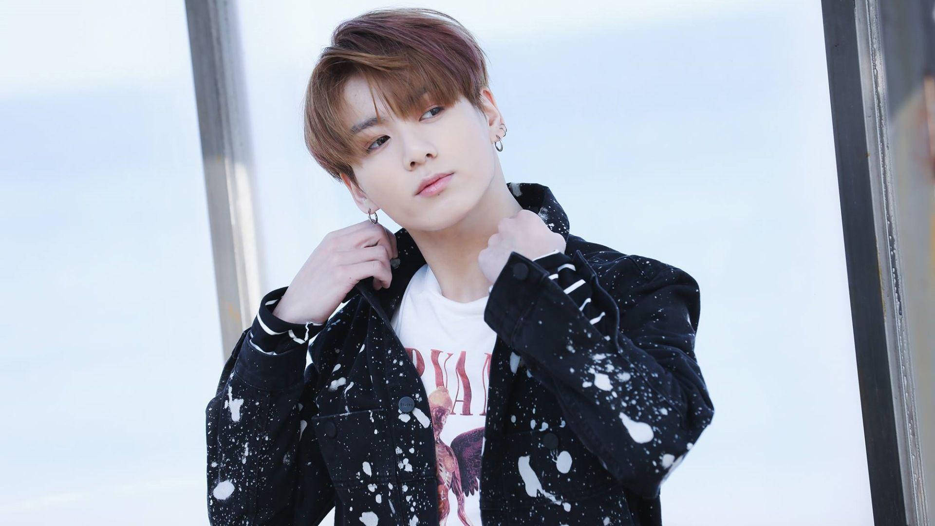 Jungkook 桌布 壁紙 — 一位年輕男子梳理著時尚髮型，在明亮的窗戶背景下調整他濺有污漬的黑色外套。