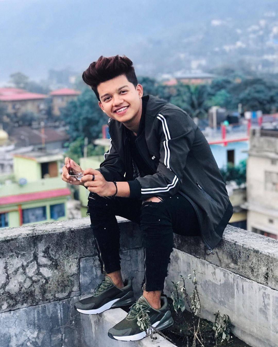 Famous 桌布 壁紙 — 一位年輕男子梳著時尚髮型，坐在屋頂邊緣微笑，手持電話，背後是城市天際線。