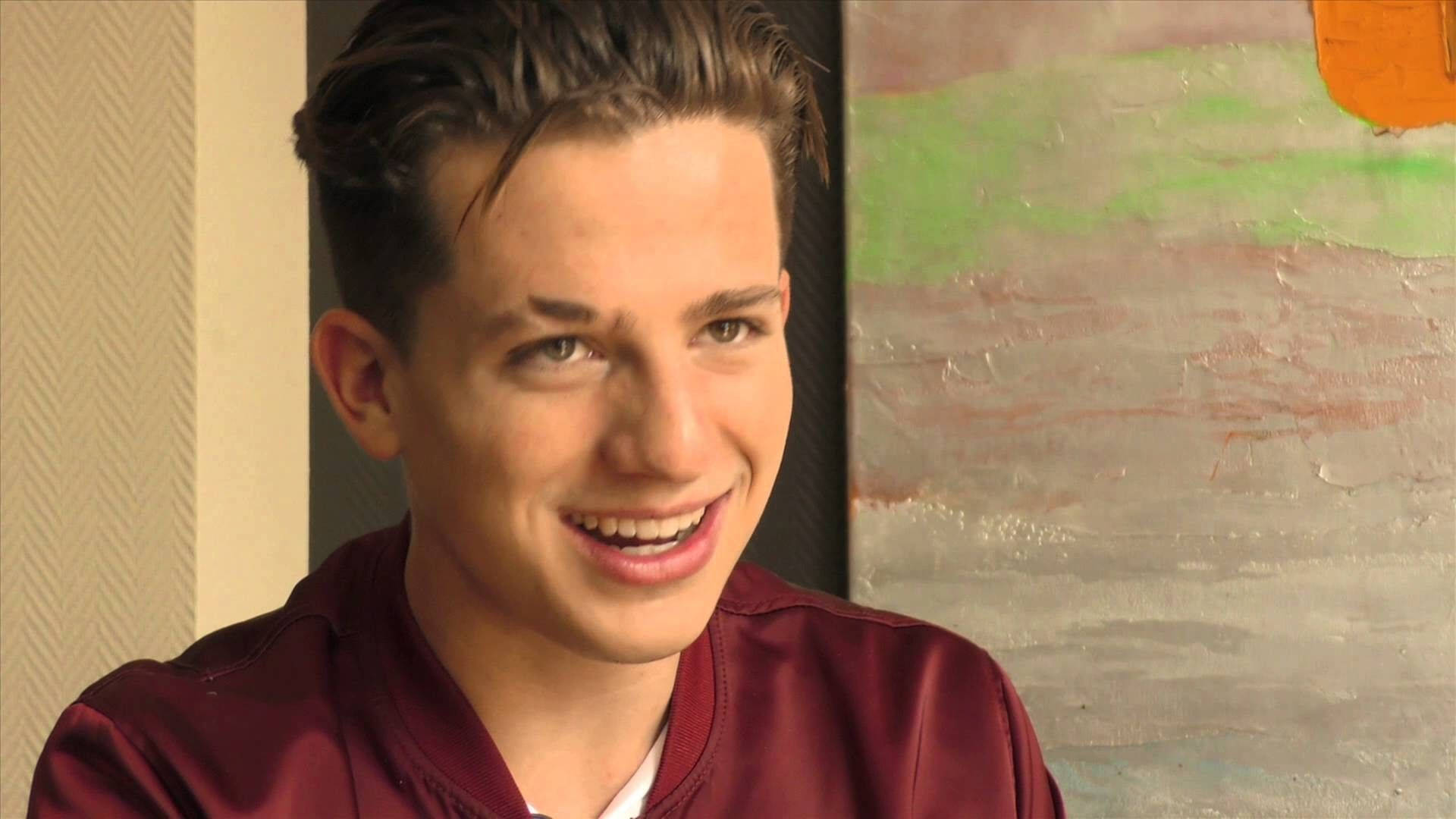 Charlie Puth 桌布 壁紙 — 一位年輕男子梳著時尚髮型，穿著栗色外套，在室內抽象藝術品附近溫暖地微笑。
