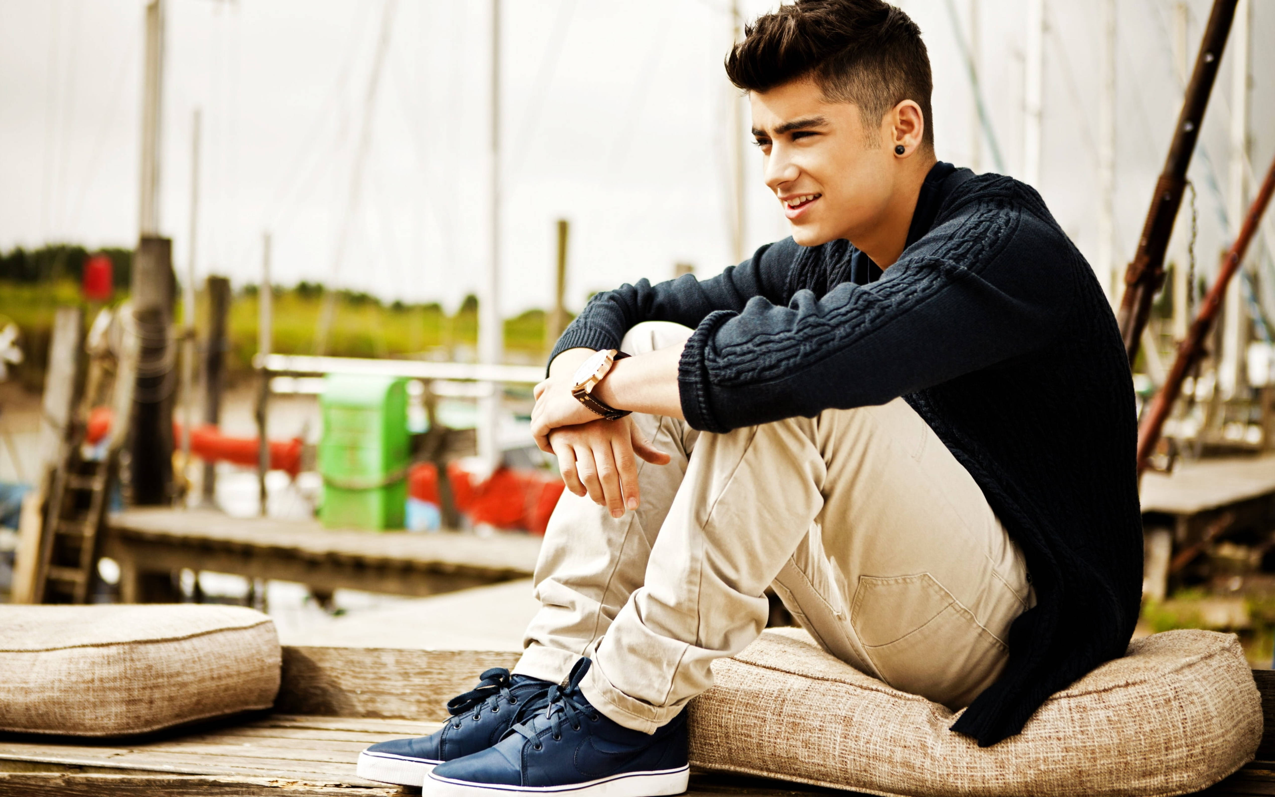 zayn 桌布 壁紙 — 一位年輕男子梳著時尚髮型，穿著深色毛衣和藍色運動鞋，隨意坐在碼頭坐墊上微笑。