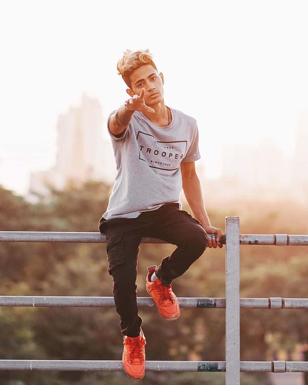 boys attitude 桌布 壁紙 — 一位年輕男子梳著時尚髮型，自信地坐在金屬欄杆上，穿著亮橙色運動鞋，背景是黃金時刻朦朧的城市天際線。