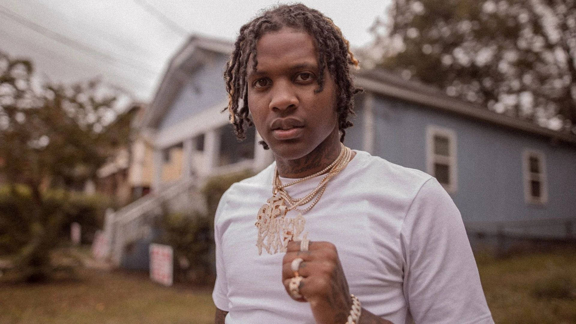 Lil Durk 桌布 壁紙 — 一位年輕男子梳著辮子、戴著金鏈，在陰天自信地站在藍色房子前。