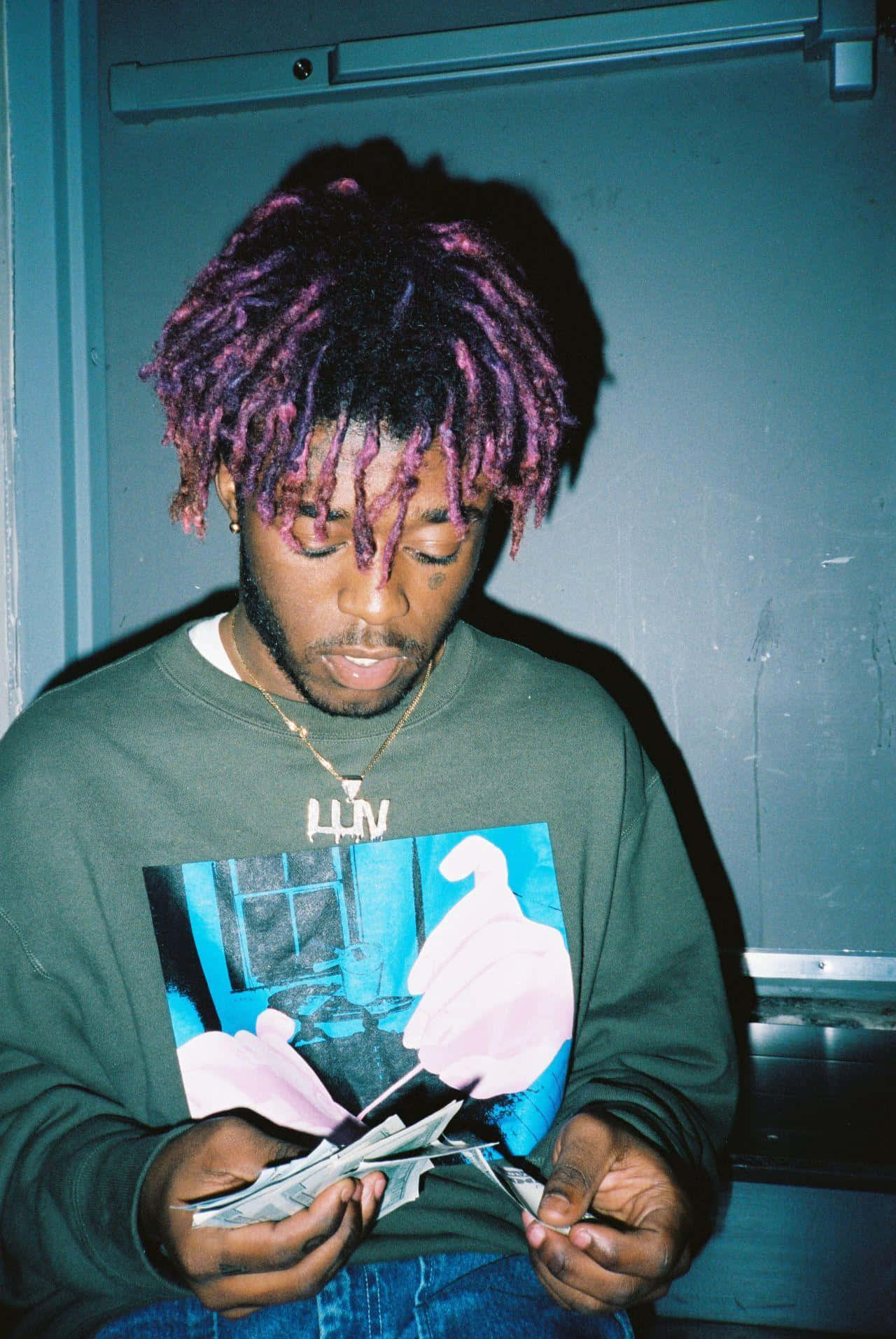 Rappers 桌布 壁紙 — 一位年輕男子留著充滿活力的紫色辮子，在燈光昏暗的房間裡穿著圖案衛衣數錢。