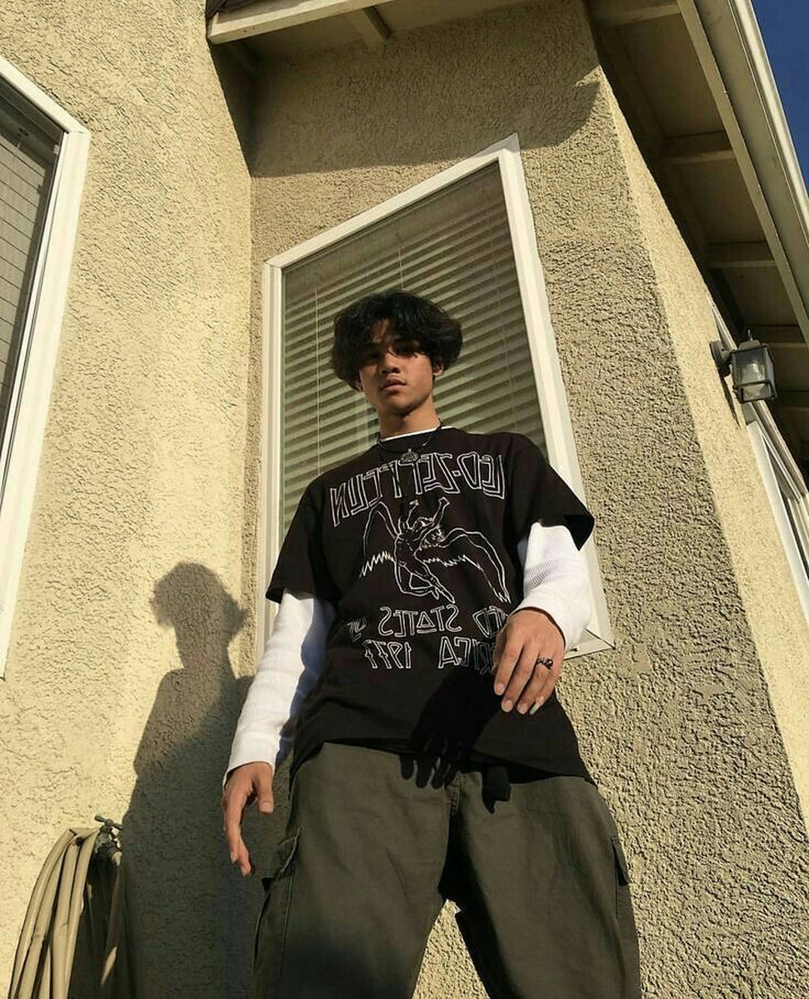 滑板男孩風格 桌布 壁紙 — 一位年輕男子留著捲髮，自信地站在米色灰泥牆前，沐浴在溫暖的金色陽光下，投下長長的影子。