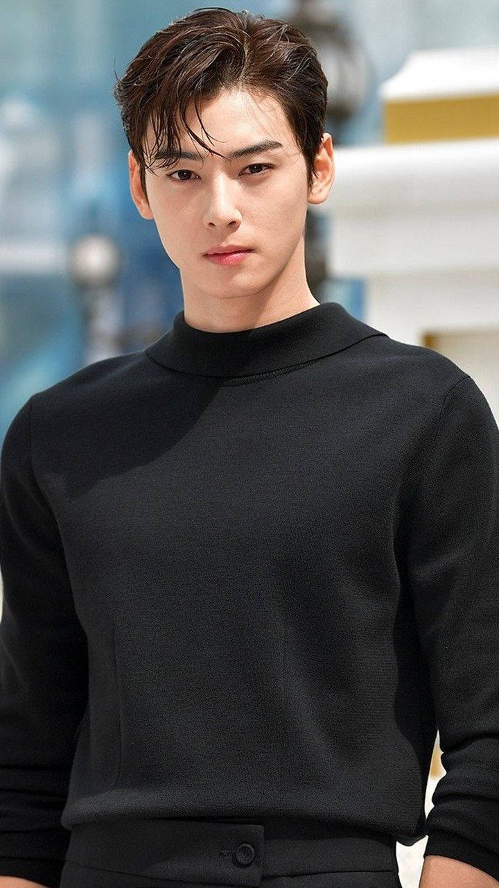 Eunwoo 桌布 壁紙 — 一位年輕男子留著時尚深色頭髮，穿著黑色高領毛衣，背景是模糊的現代建築。
