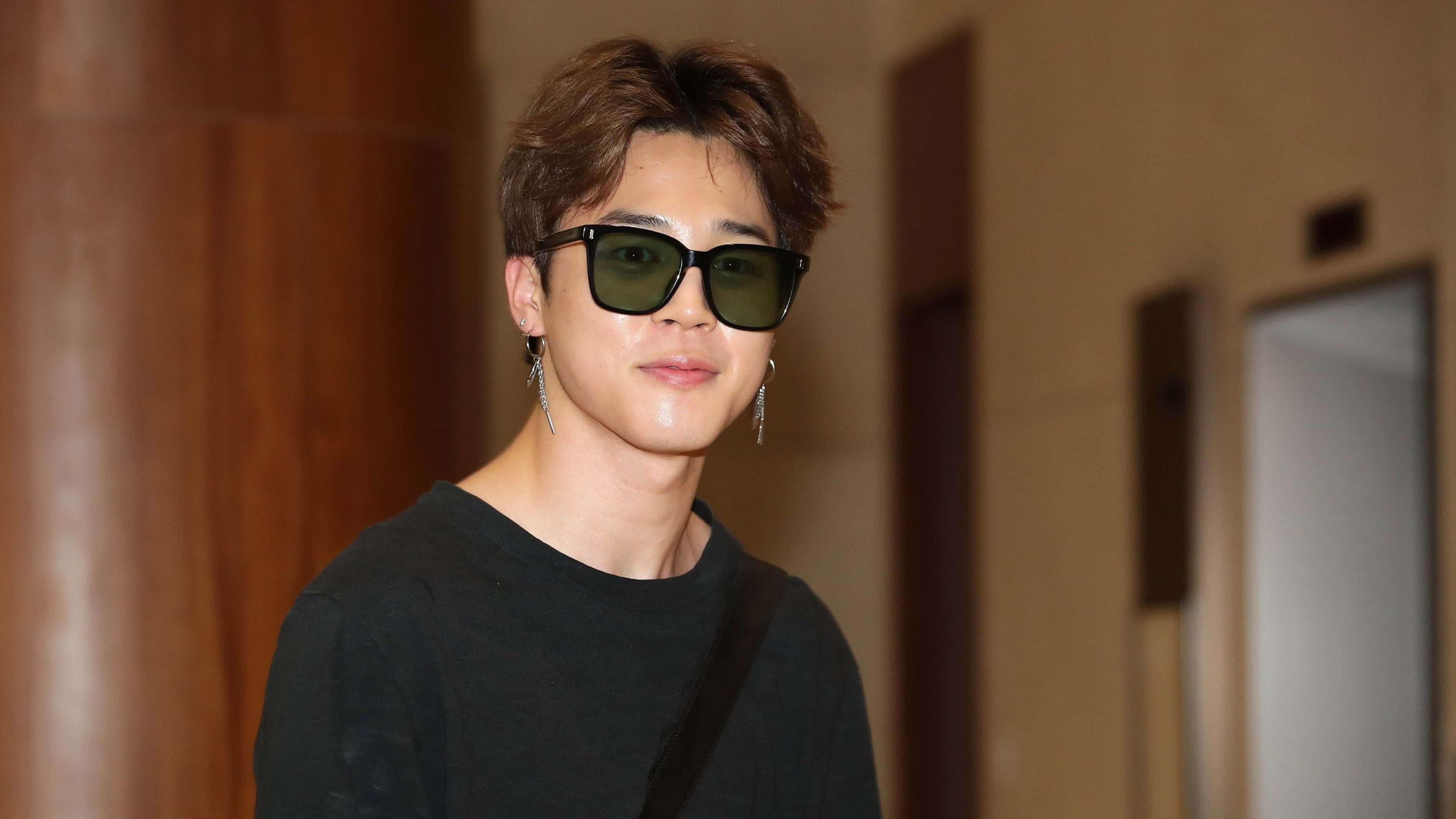 BTS 桌布 壁紙 — 一位年輕男子留著時尚髮型，戴著深色太陽眼鏡和垂墜耳環，站在室內的走廊裡。