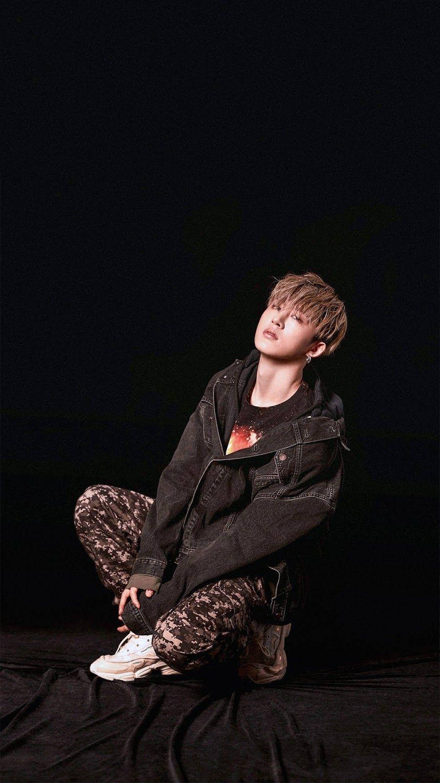 kim hanbin 桌布 壁紙 — 一位年輕男子留著時尚髮型，盤腿坐在黑色布料上，背景昏暗，身穿牛仔外套和圖案褲子。