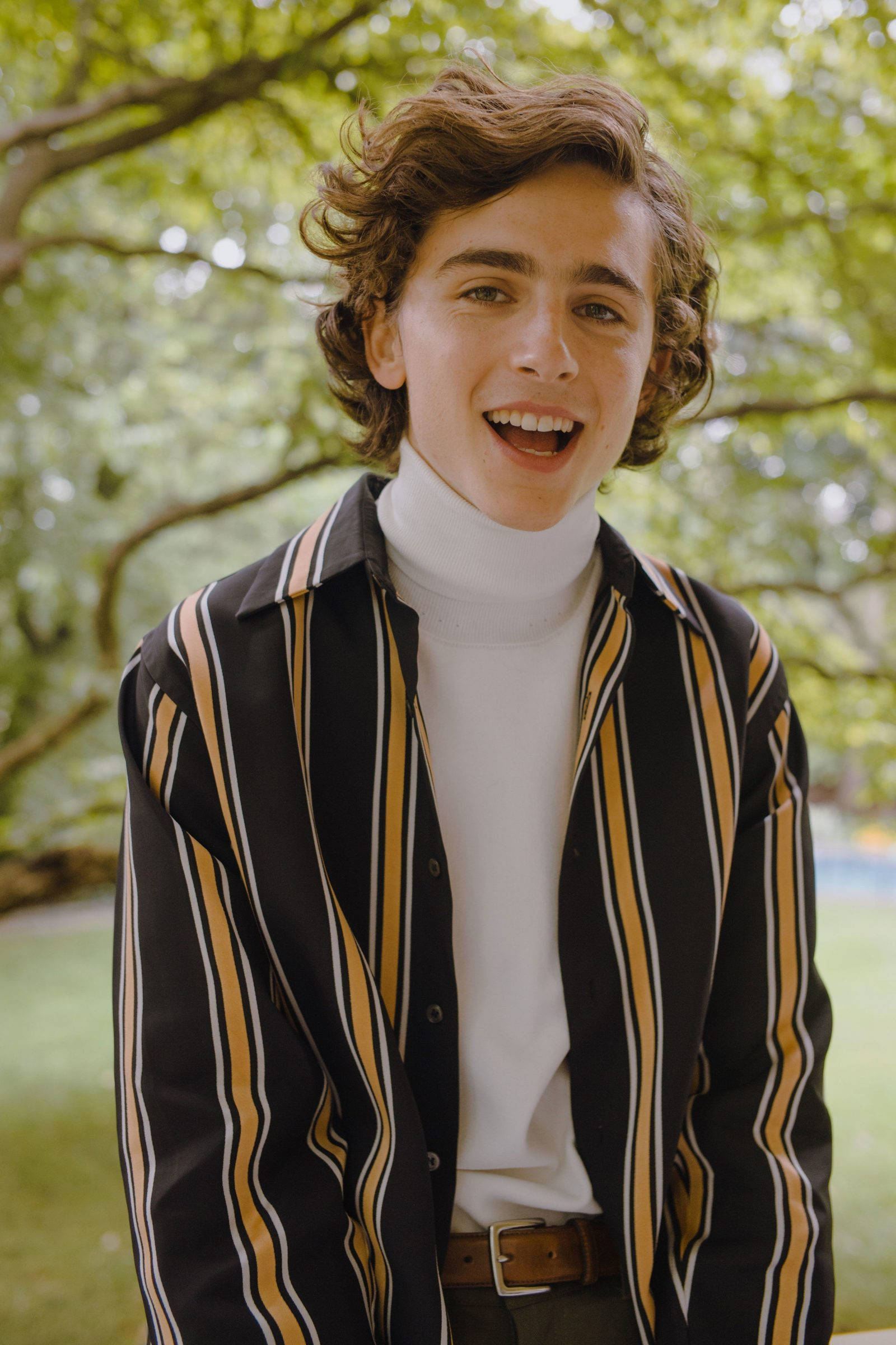 Timothée Chalamet 桌布 壁紙 — 一位年輕男子留著棕色捲髮，在戶外穿著條紋襯衫內搭白色高領毛衣，背景是茂密的綠樹，笑容燦爛。