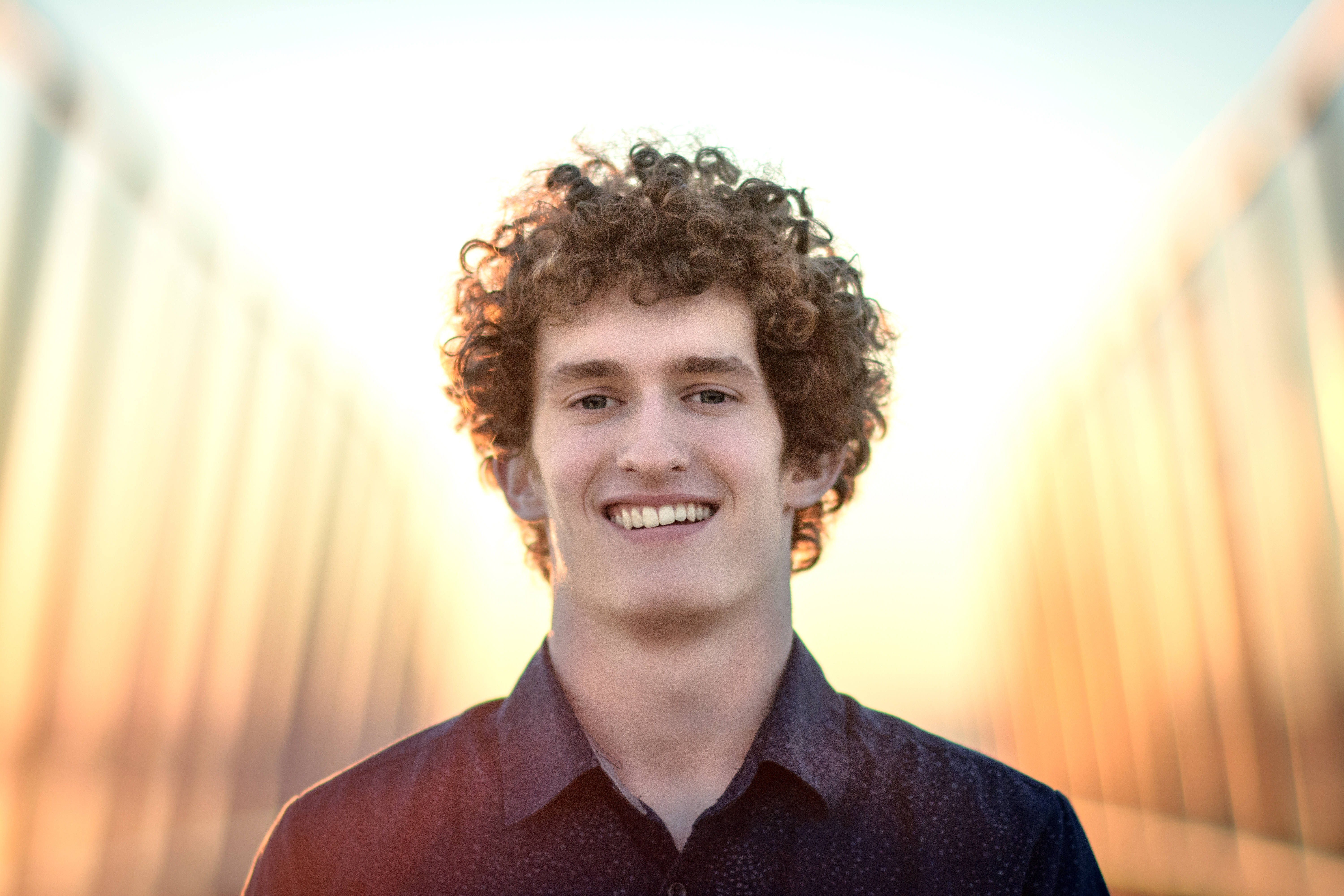 smiling 桌布 壁紙 — 一位年輕男子留著棕色捲髮，在柔和模糊的金色日落背景下溫暖地微笑。