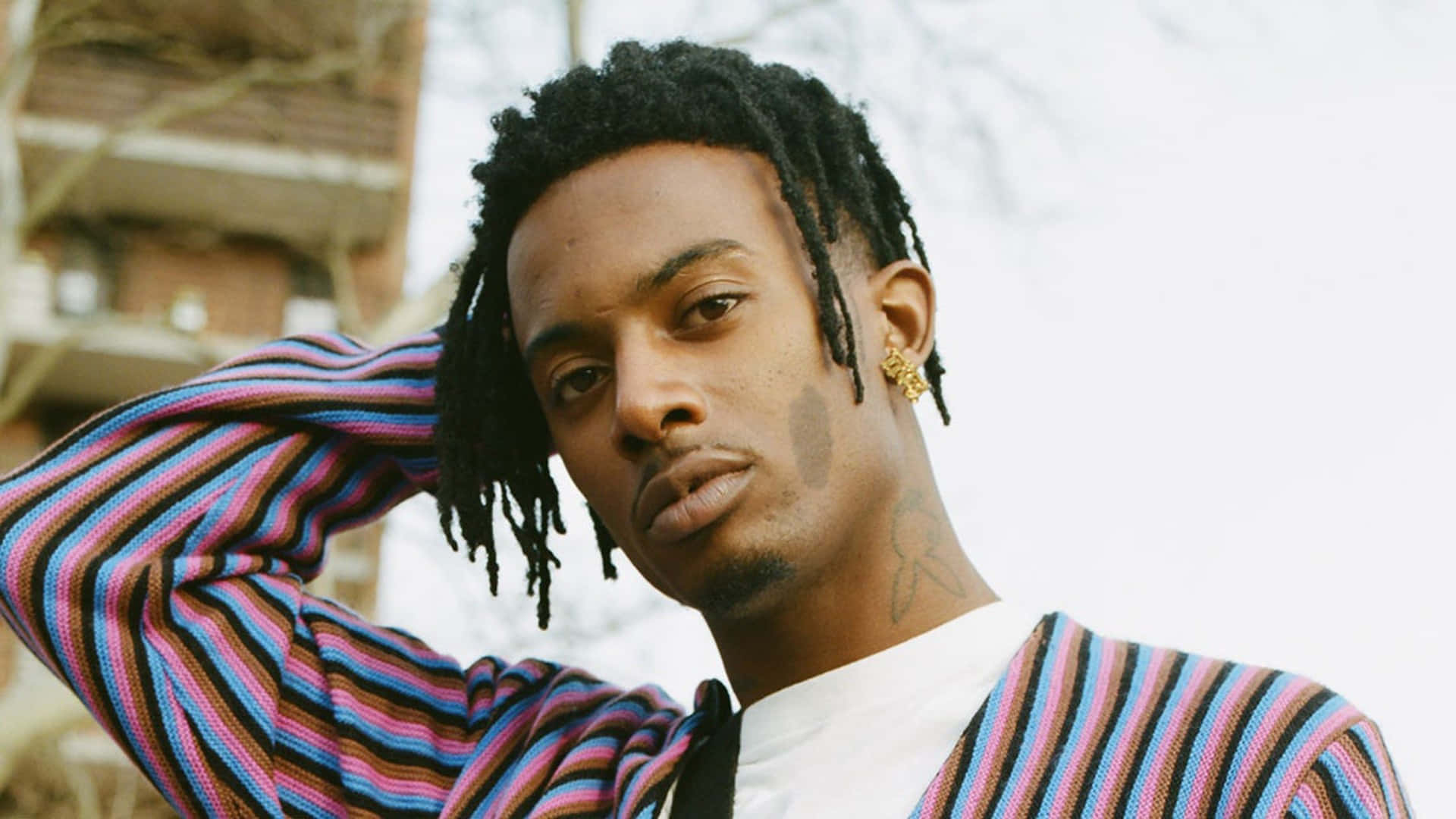 Playboi Carti HD 桌布 壁紙 — 一位年輕男子留著辮子頭、戴著金色耳環，穿著色彩鮮豔的條紋毛衣，在模糊的城市背景下自信地擺姿勢。