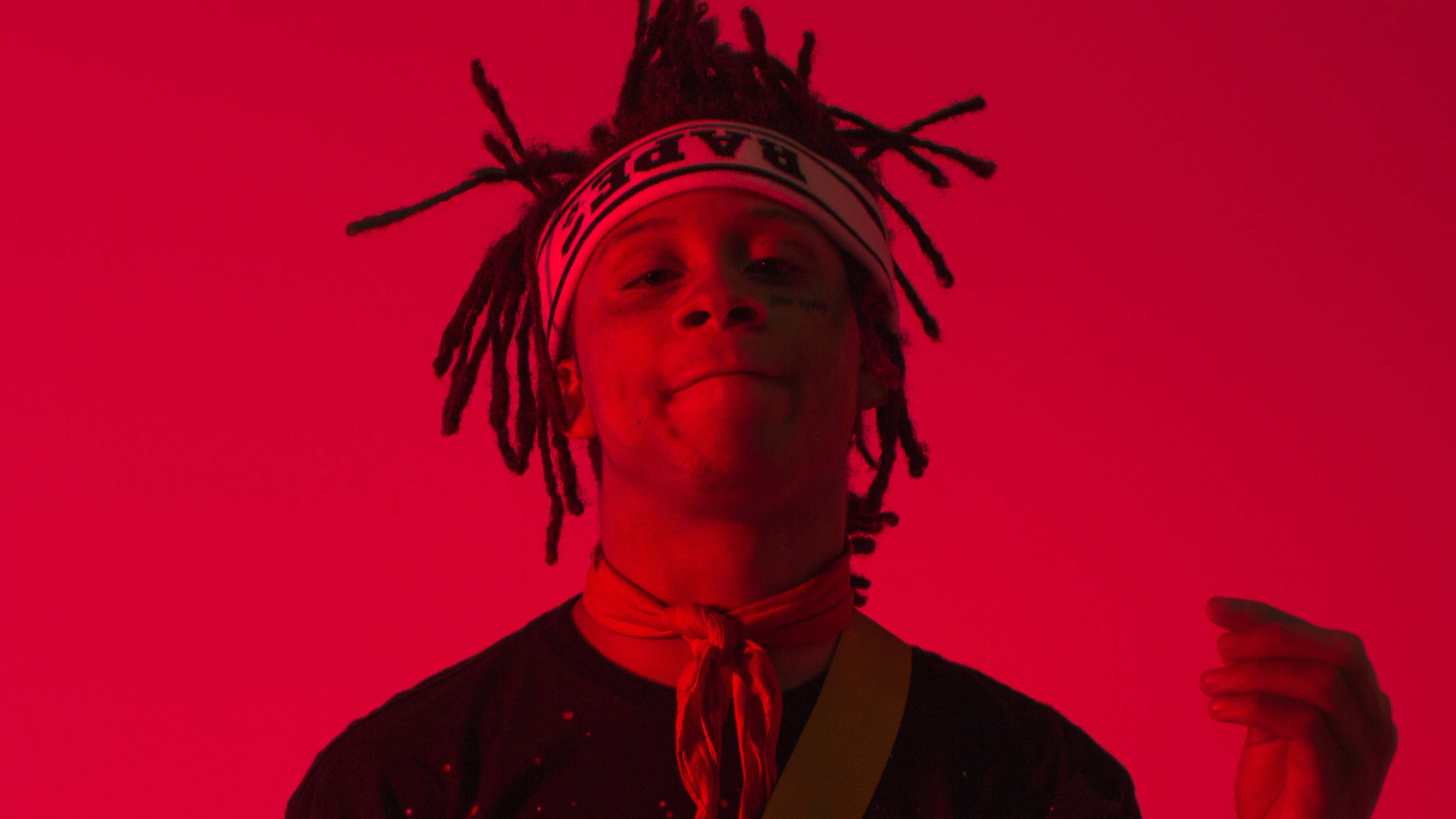 Trippie Redd 桌布 壁紙 — 一位年輕男子留著辮子頭、戴著頭帶，在鮮紅色的攝影棚背景前溫柔微笑。