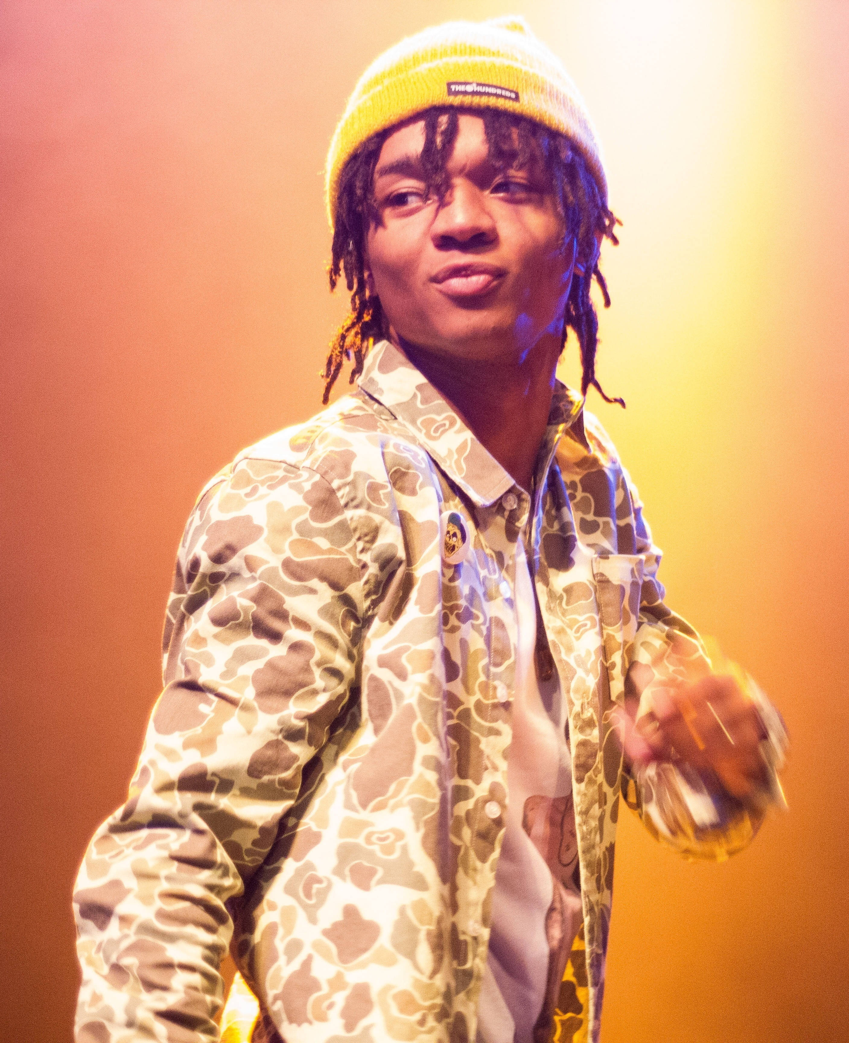 Swae Lee 桌布 壁紙 — 一位年輕男子留著辮子頭，戴著黃色無邊帽，身穿迷彩外套，在溫暖的橙黃燈光下於舞台上表演。