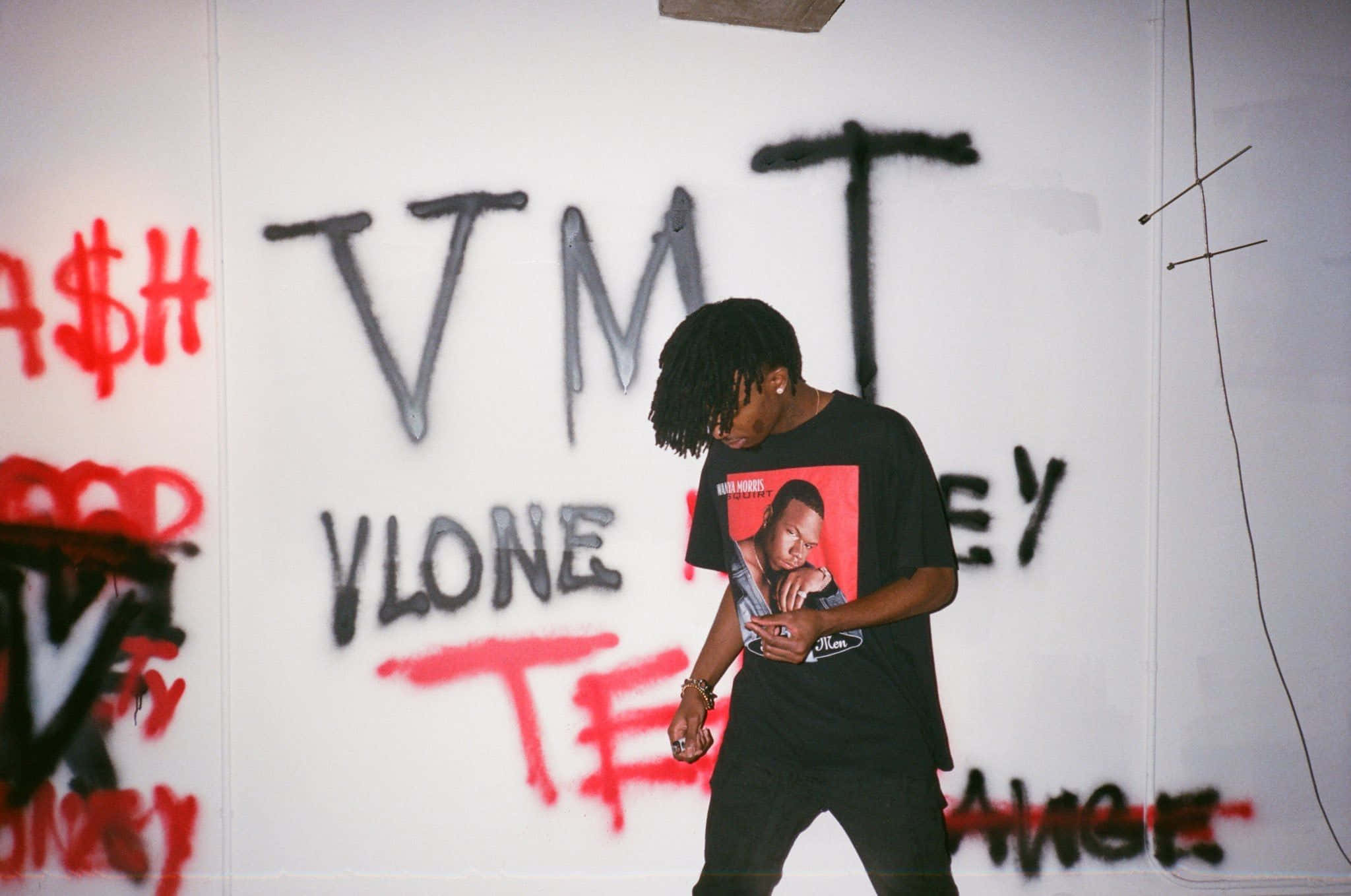 Playboi Carti 桌布 壁紙 — 一位年輕男子留著辮子頭，站在一個佈滿塗鴉的房間裡，身穿帶有紅色圖案的黑色T恤。