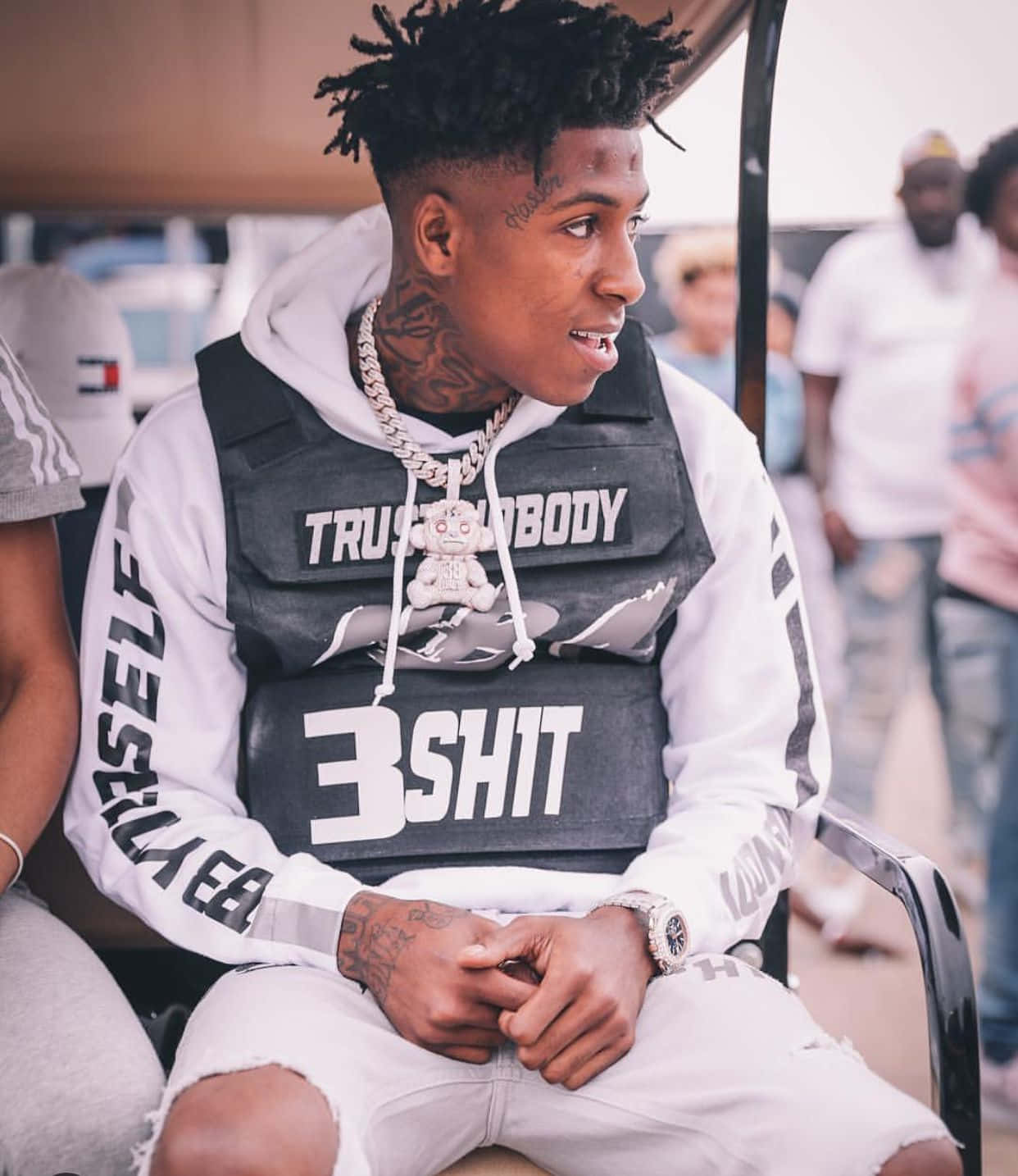 NBA YoungBoy 桌布 壁紙 — 一位年輕男子留著辮子頭和刺青，穿著黑色背心，內搭印有粗體文字的白色連帽衫，坐在戶外。