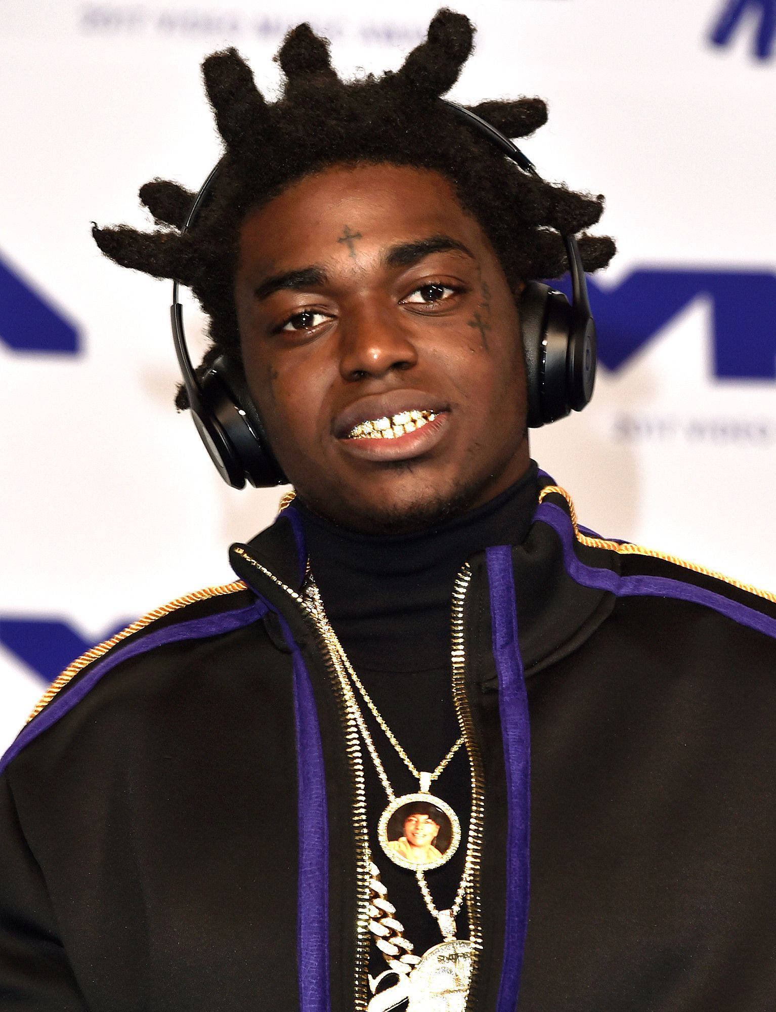 Kodak 桌布 壁紙 — 一位年輕男子留著辮子頭和金色牙齒，戴著耳機和多層項鍊，背景為品牌背景。
