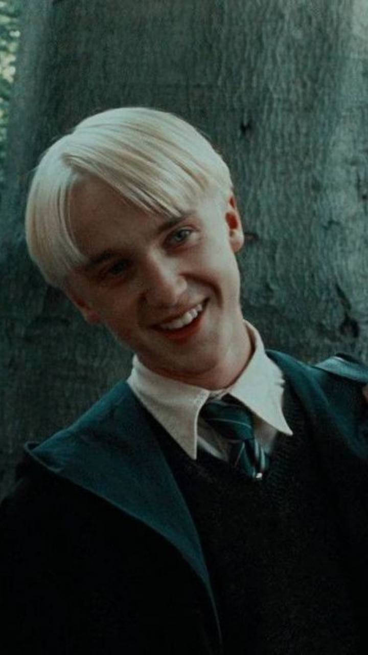 史萊哲林 桌布 壁紙 — 一位年輕男子留著鉑金色頭髮，在昏暗的森林光線中，靠著紋理分明的樹幹溫暖微笑。