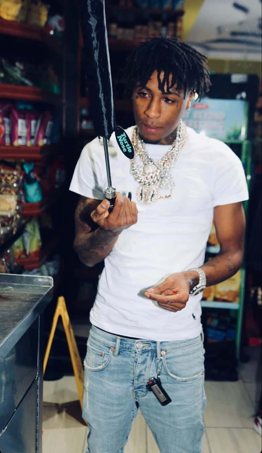 Youngboy 桌布 壁紙 — 一位年輕男子留著雷鬼辮，戴著鑽石鏈，在燈光明亮的便利商店裡撐著一把黑色雨傘。