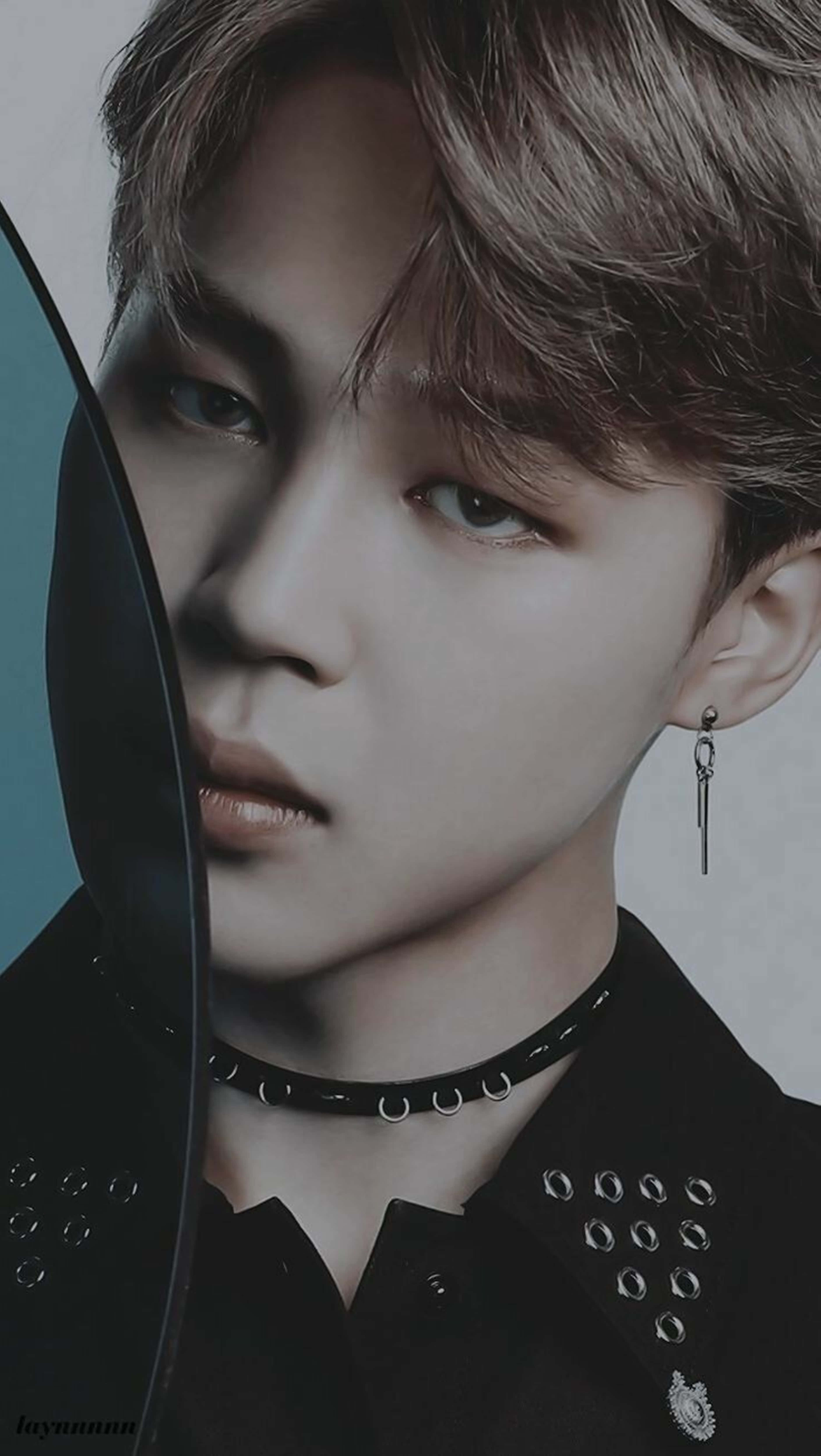 Bts Jimin 桌布 壁紙 — 一位年輕男子的特寫肖像，淺棕色頭髮，眼神銳利，身穿黑色鉚釘襯衫和頸鏈，手持一張黑色黑膠唱片，部分遮擋臉部，背景為柔和的青綠色和灰色。