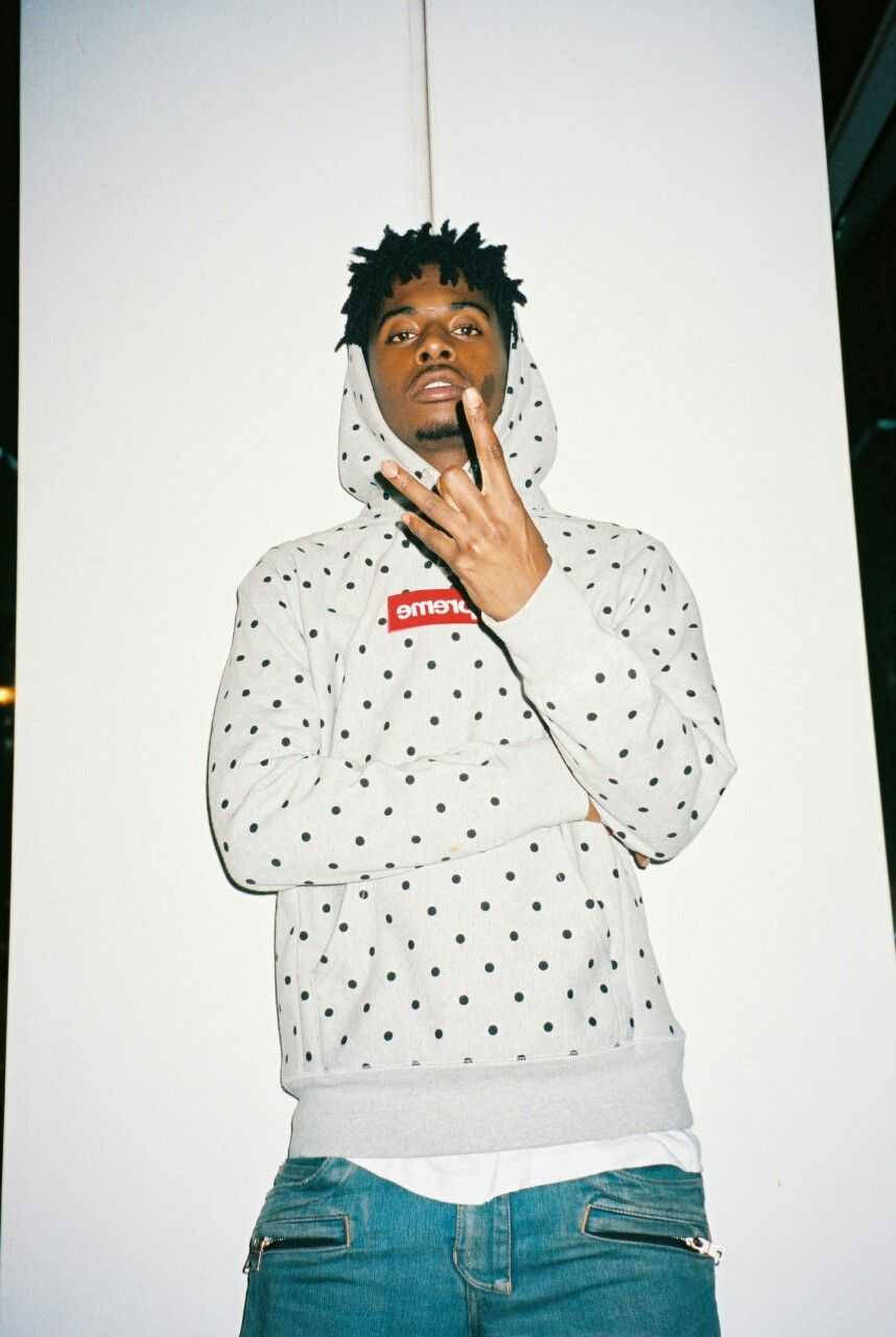 Carti 桌布 壁紙 — 一位年輕男子穿著圓點連帽衫，自信地靠著白牆擺姿勢，一隻手靠近臉部。