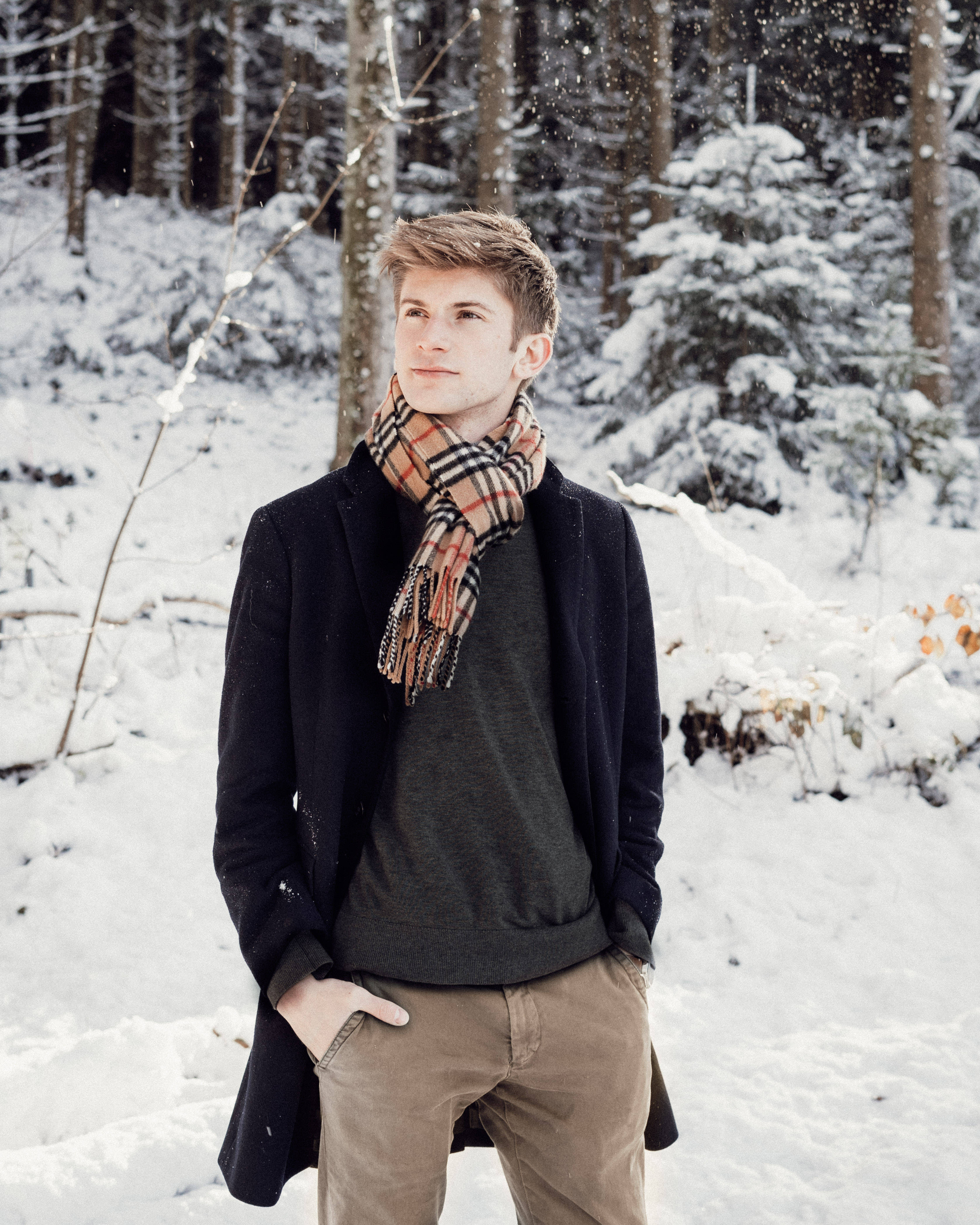 Burberry 桌布 壁紙 — 一位年輕男子穿著格紋圍巾和大衣，靜靜地站在雪地森林中，雪花輕輕飄落在他周圍。