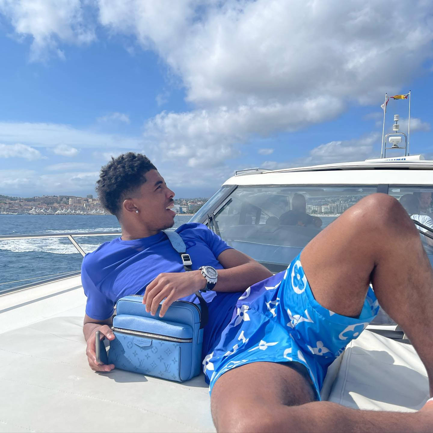 Yacht 桌布 壁紙 — 一位年輕男子穿著藍色泳裝，拿著相配的袋子，在晴朗天空和零星雲朵下，隨意地靠在遊艇甲板上。