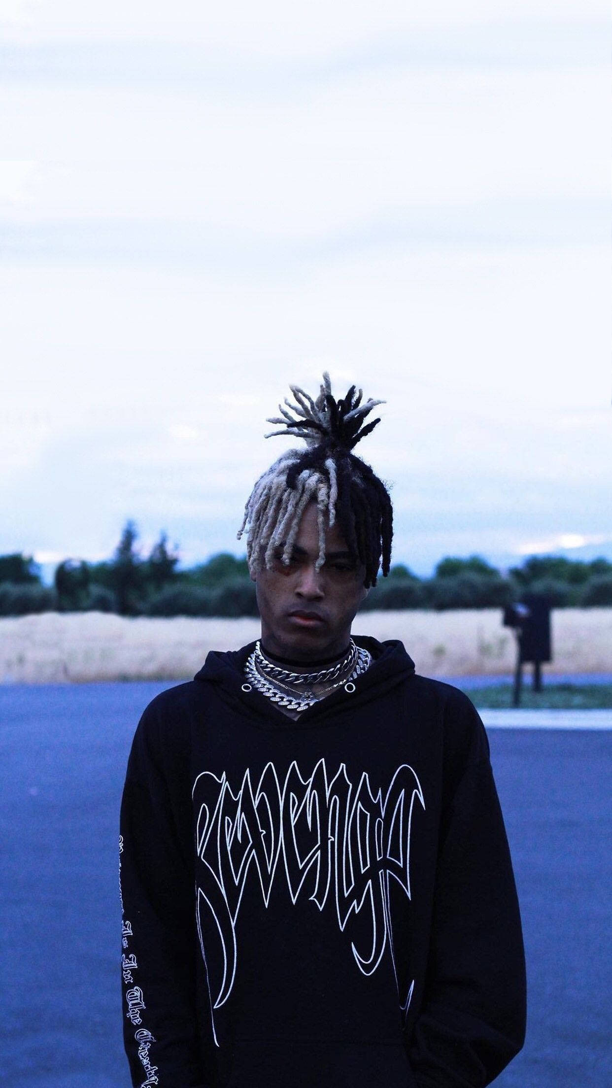 XXXTENTACION 桌布 壁紙 — 一位年輕男子編著辮子，戴著多層鏈條，在陰天、冷色調的戶外環境中自信地站立。