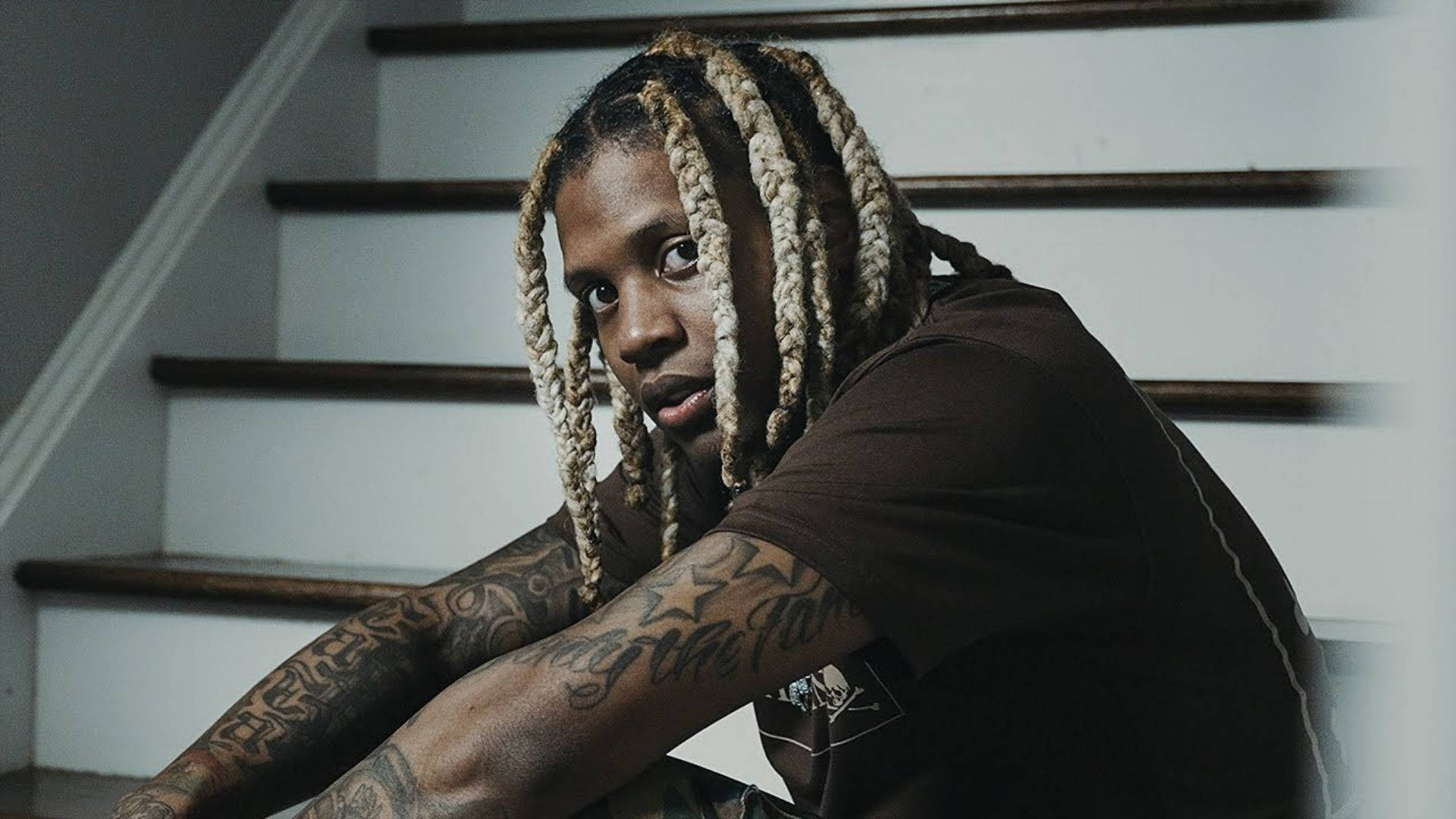 Lil Durk 桌布 壁紙 — 一位年輕男子編著辮子、身上有刺青，坐在樓梯上，專注地凝視鏡頭。