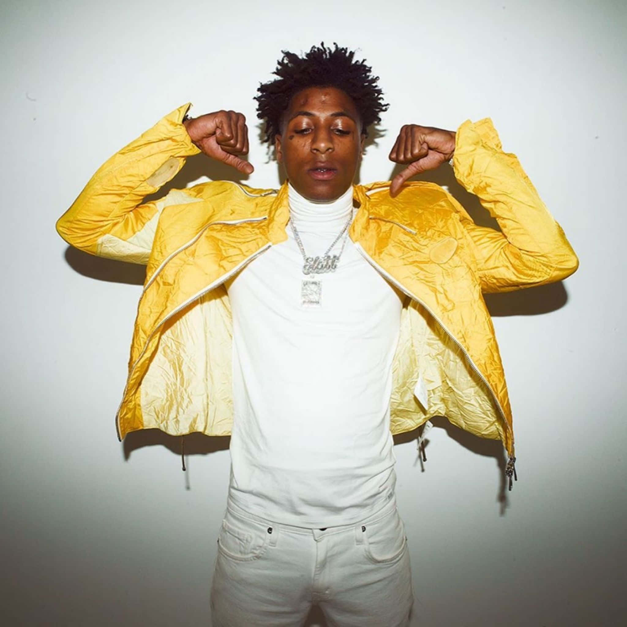 NBA YoungBoy 桌布 壁紙 — 一位年輕男子身穿亮黃色外套和白色高領毛衣，在純白牆壁前自信地舉起雙臂擺姿勢。