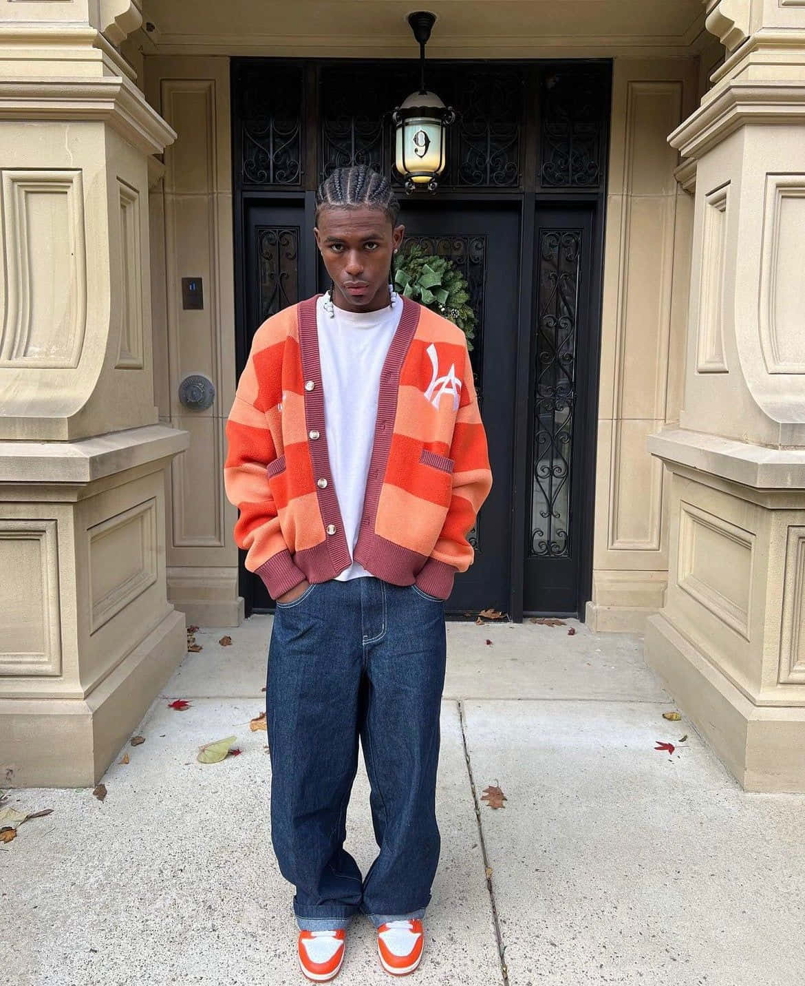 vetements 桌布 壁紙 — 一位年輕男子身穿大膽的橙色條紋開襟衫和藍色牛仔褲，自信地站在優雅米色豪宅入口前的人行道上。