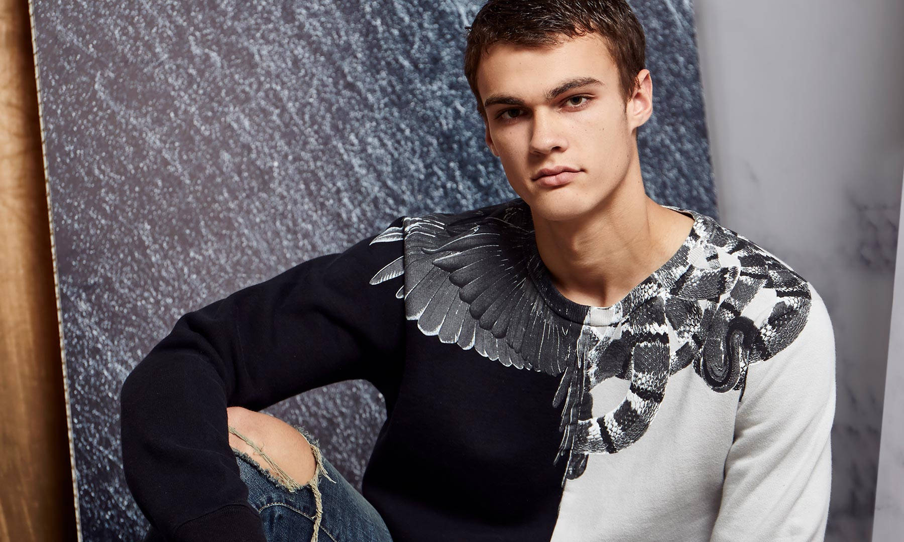 Fashion Model 桌布 壁紙 — 一位年輕男子身穿帶有翅膀和蛇圖案的黑白圖案毛衣，在紋理灰色背景前擺姿勢。
