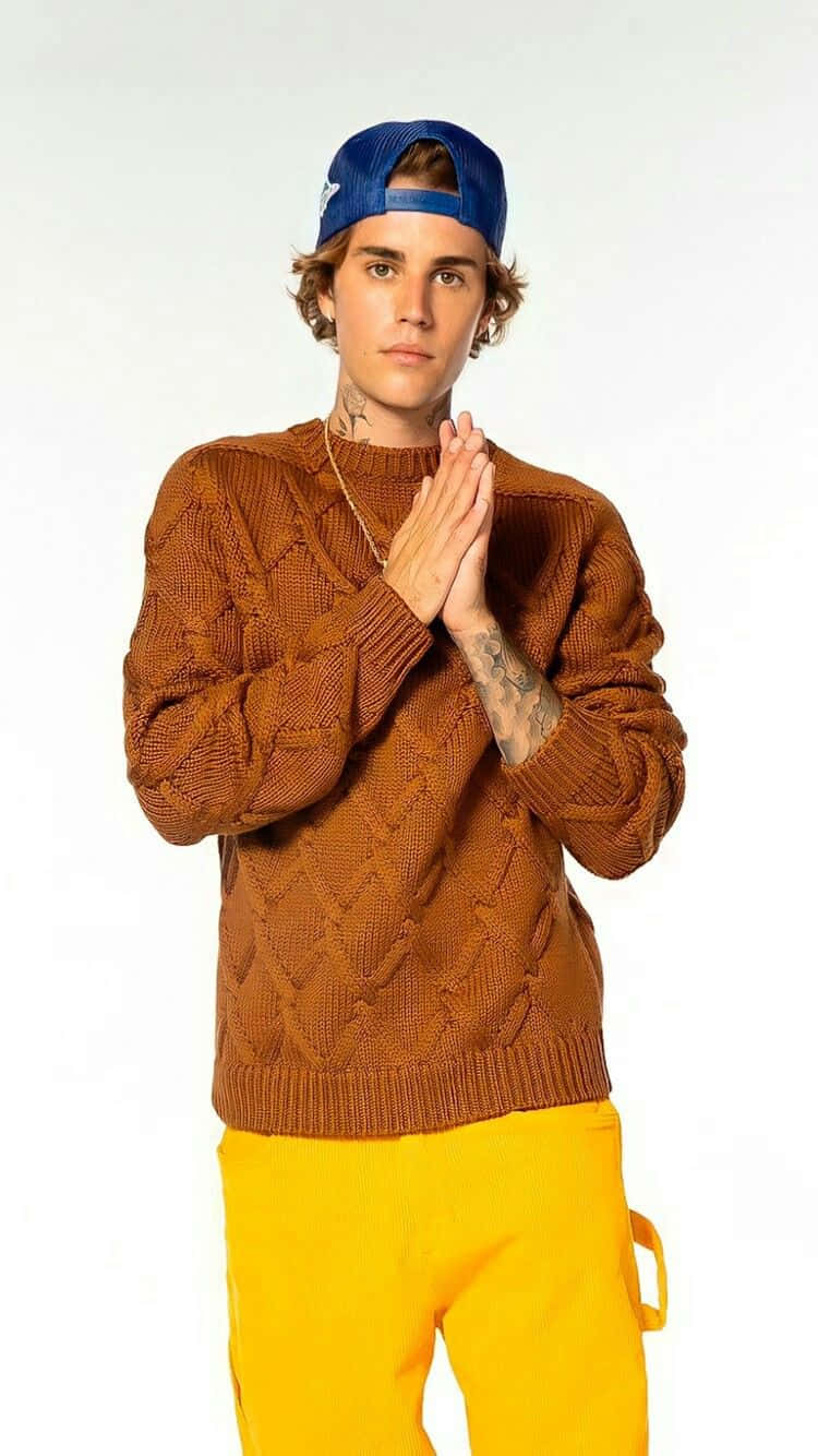 Justin Bieber 2021 桌布 壁紙 — 一位年輕男子身穿棕色麻花針織毛衣和黃色褲子，雙手交握，在純白背景前擺姿勢。