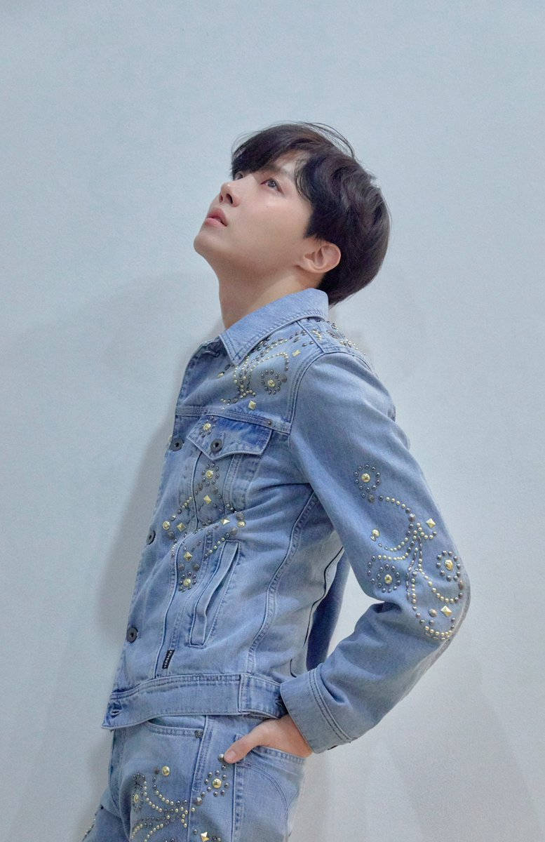 j-hope 桌布 壁紙 — 一位年輕男子身穿淺藍色牛仔外套和褲子，飾有金色鉚釘，抬頭望著純灰色的牆壁。