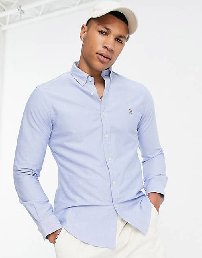 ralph lauren 桌布 壁紙 — 一位年輕男子身穿淺藍色鈕扣襯衫和白色帽子，雙手插袋，在明亮的攝影棚背景下自信地擺姿勢。