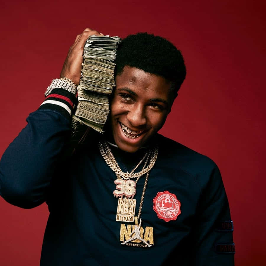 Celebrity 桌布 壁紙 — 一位年輕男子露出燦爛笑容，將一疊厚厚的現鈔貼在頭上，佩戴閃亮首飾，身穿品牌衛衣，背景為深紅色。