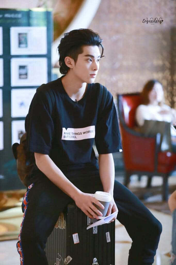 Dylan Wang 桌布 壁紙 — 一位年輕男子頭髮造型時尚，悠閒地坐在行李箱上，在溫暖的室內空間中手持咖啡杯。