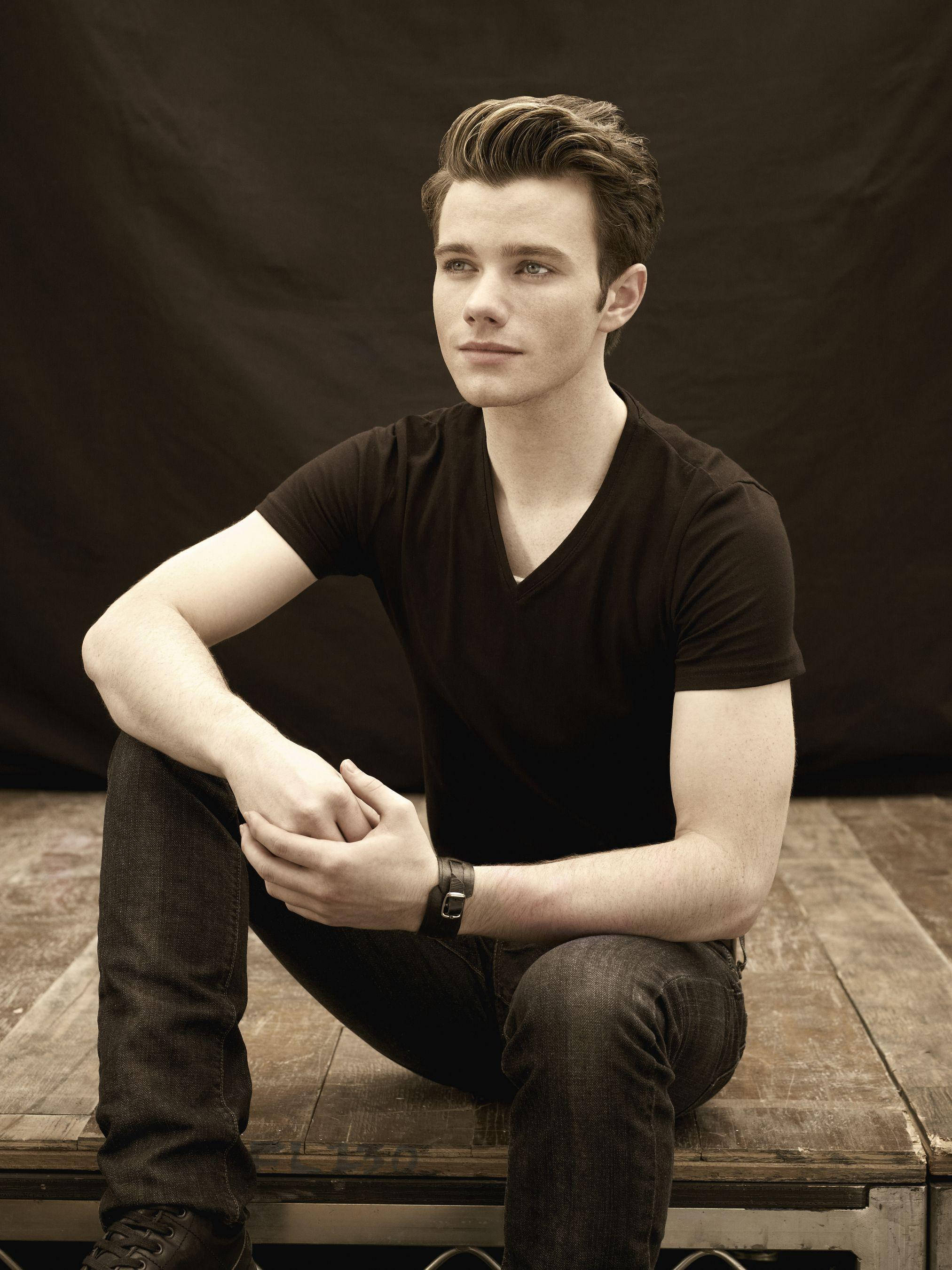Chris Colfer 桌布 壁紙 — 一位年輕男子頭髮造型時尚，隨意地坐在木長椅上，背景昏暗，身穿黑色V領襯衫和牛仔褲。