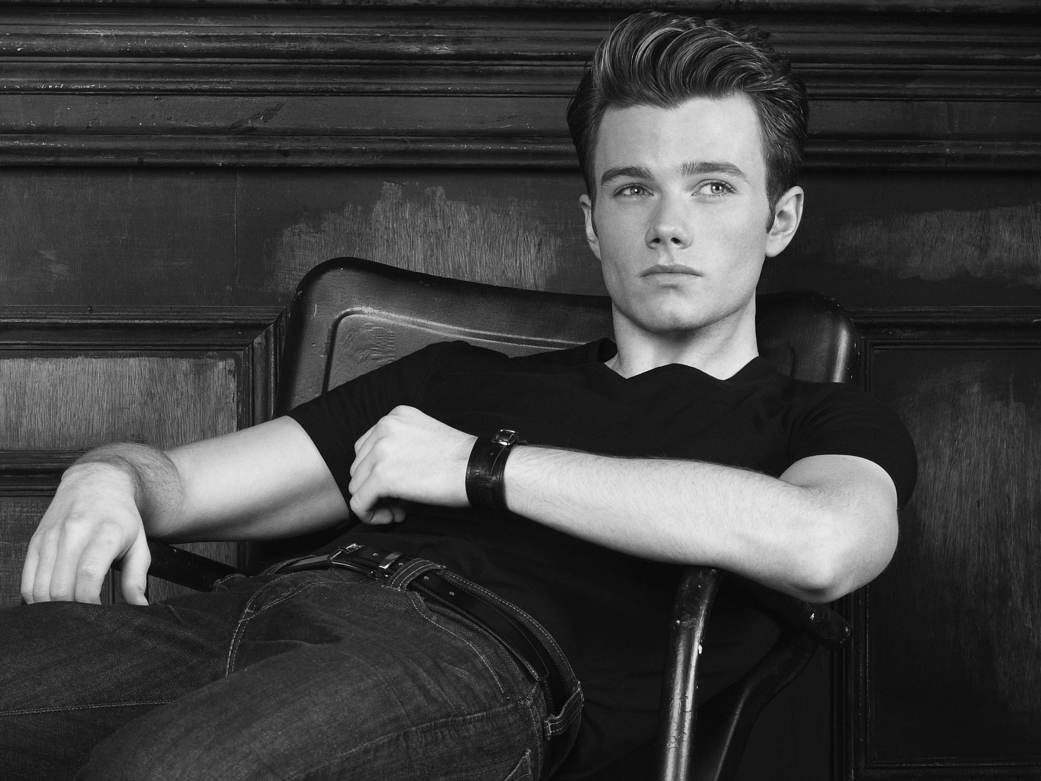 Chris Colfer 桌布 壁紙 — 一位年輕男性自信地坐在皮革椅上，背景是深色木質牆板，呈現出鮮明的黑白肖像。