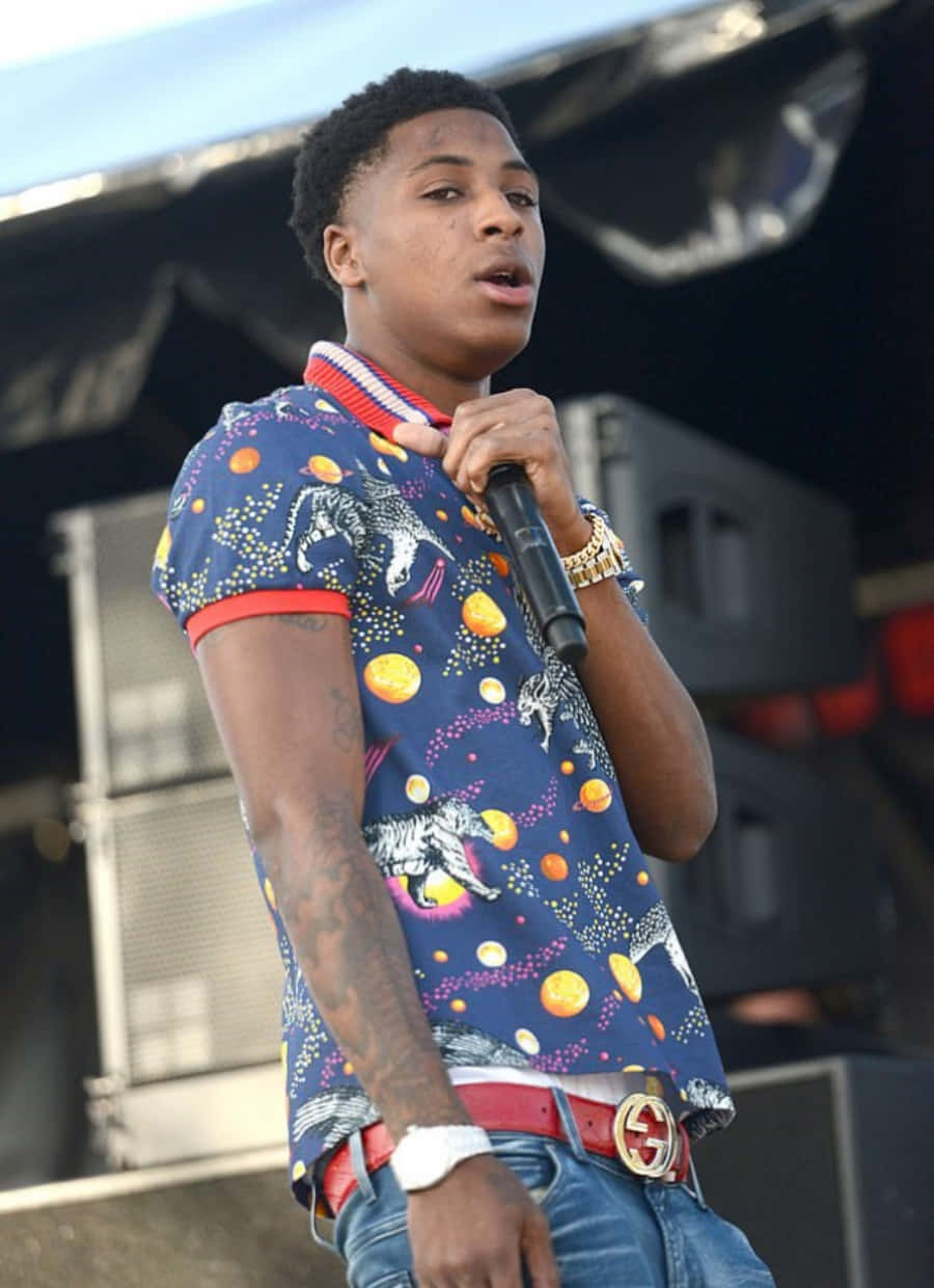 NBA YoungBoy 桌布 壁紙 — 一位年輕男性表演者在舞台上對著麥克風唱歌，身穿帶有恐龍和行星的鮮豔藍色圖案襯衫。