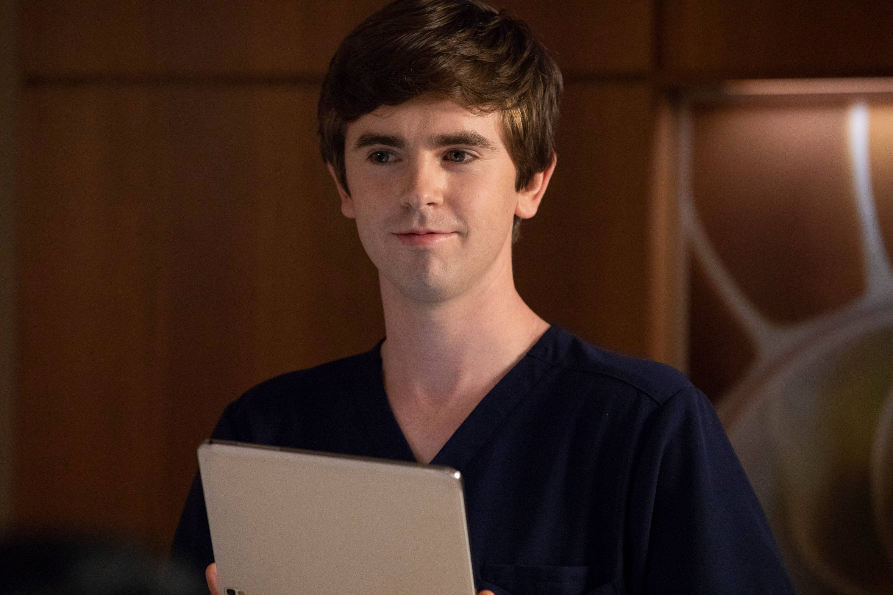 The Good Doctor 桌布 壁紙 — 一位年輕男醫生穿著海軍藍色刷手服，在溫暖的木質背景下拿著平板電腦。