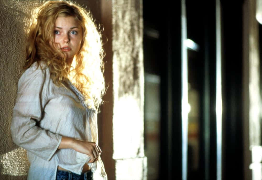 Izabella Miko 桌布 壁紙 — 一位年輕的捲曲金髮女子站在陽光照射的門口，她的臉龐在黑暗背景下被溫暖的光線照亮。